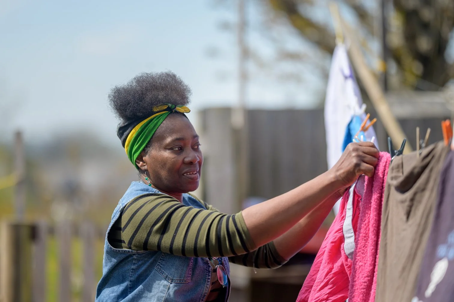 Woman hanging clothes on a clothesline outdoors, wearing a colorful headscarf and a denim vest.