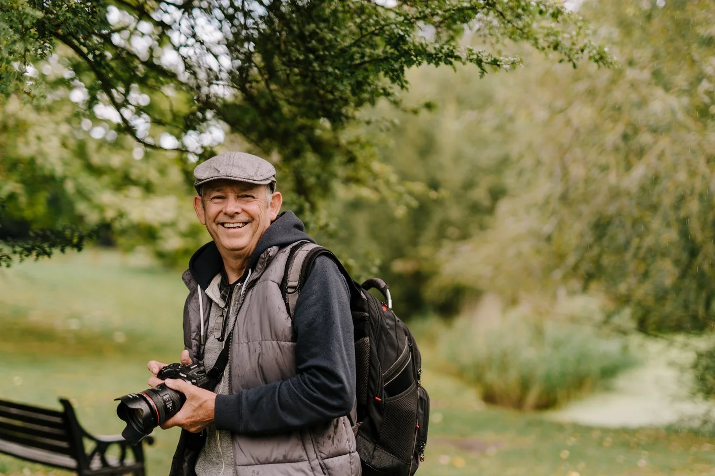 A smiling man wearing a gray cap, dark hoodie, and gray vest, holding a camera. He has a backpack and is standing outdoors in a park with green trees and a bench in the background.