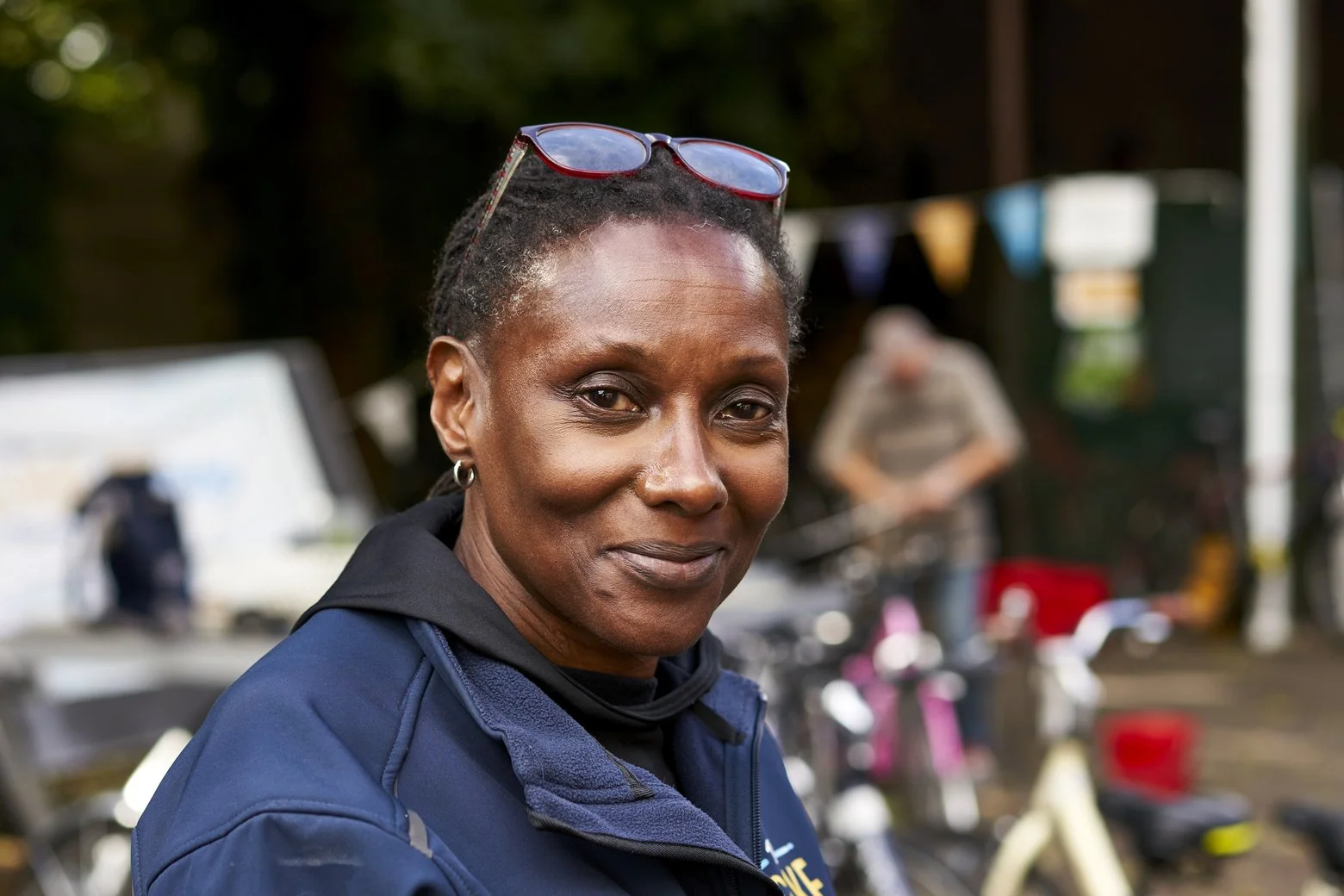 A woman with short, curly hair, wearing glasses on her head, earrings, and a navy jacket, smiling outdoors with blurred bicycles and a person in the background.