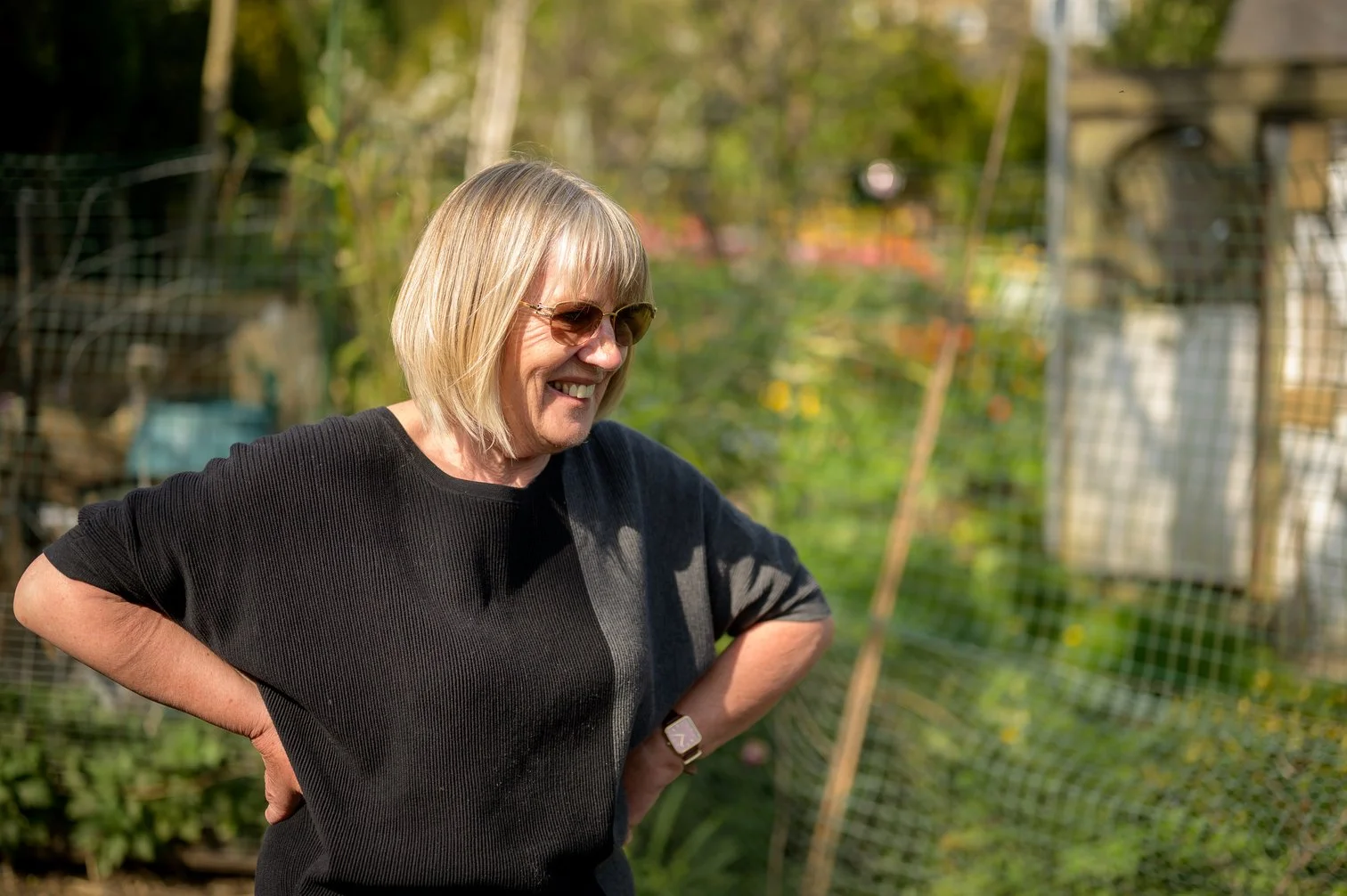 A woman with blonde hair, wearing sunglasses and a black shirt, smiling outdoors in a garden or farm setting.