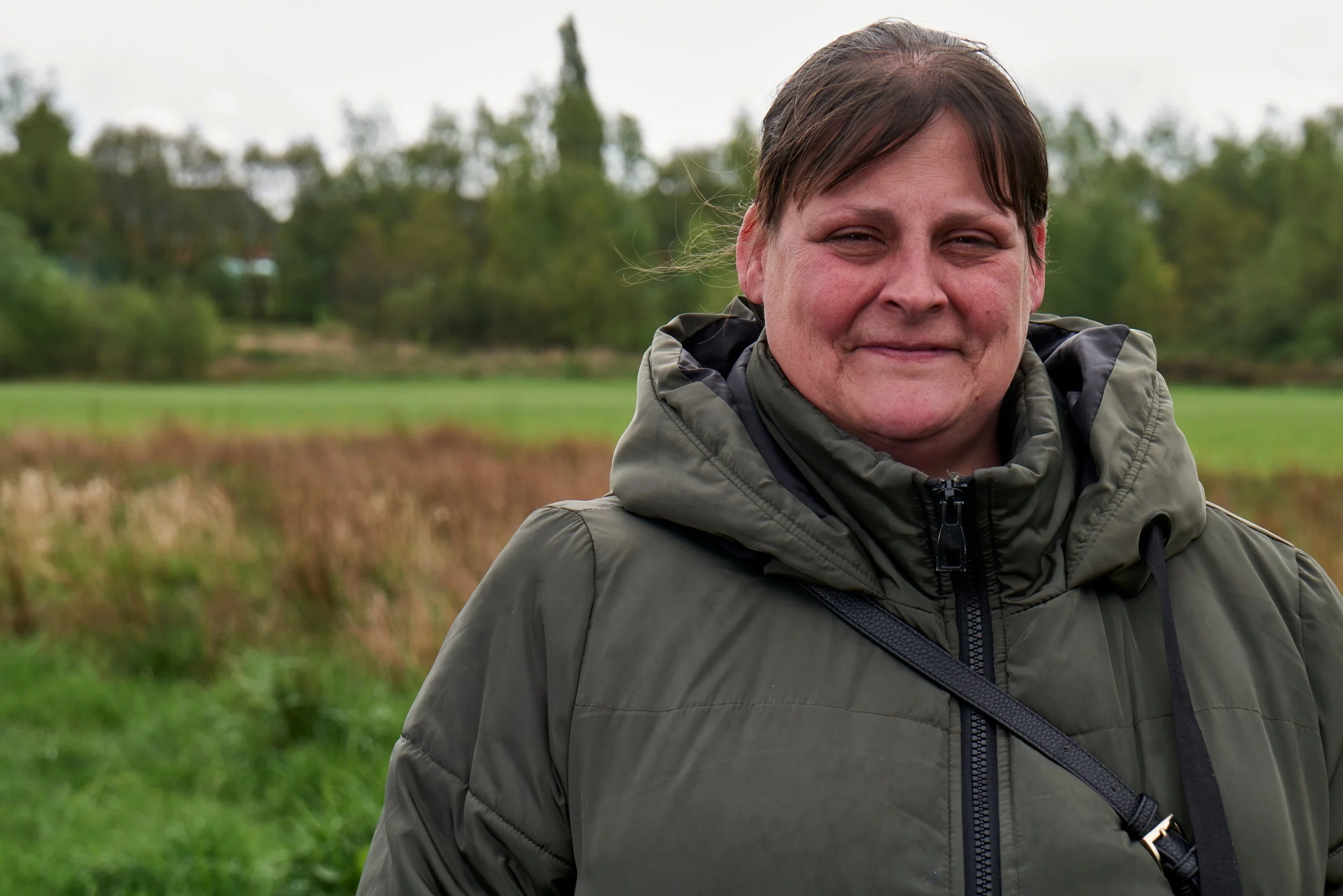 A person with short dark hair smiling outdoors in a green field with trees in the background, wearing a dark green puffer jacket.