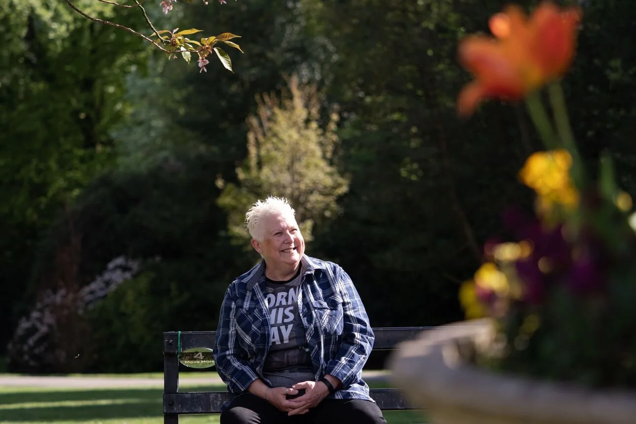 An elderly woman with short white hair sitting on a park bench, smiling and looking to her left, wearing a plaid shirt over a black t-shirt with white text, in a park with green trees and a blurred flower arrangement in the foreground.