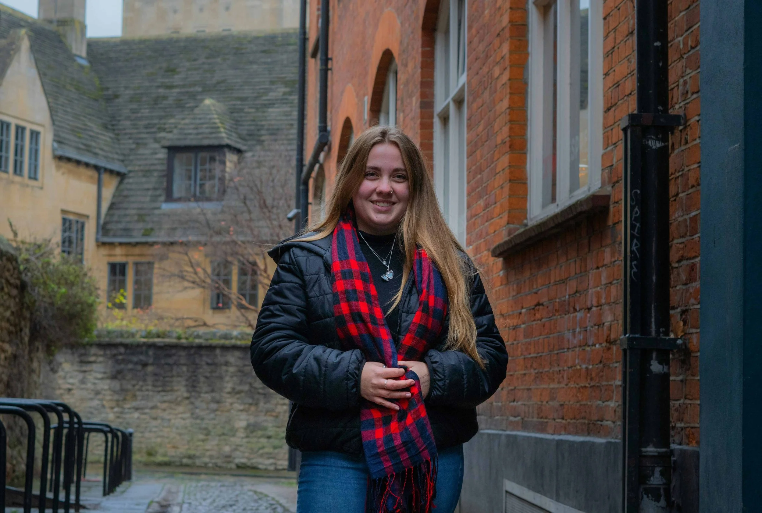 A young woman with long blonde hair smiling, wearing a black jacket and a red and blue checkered scarf, standing on a narrow cobblestone alley between brick and stone buildings.
