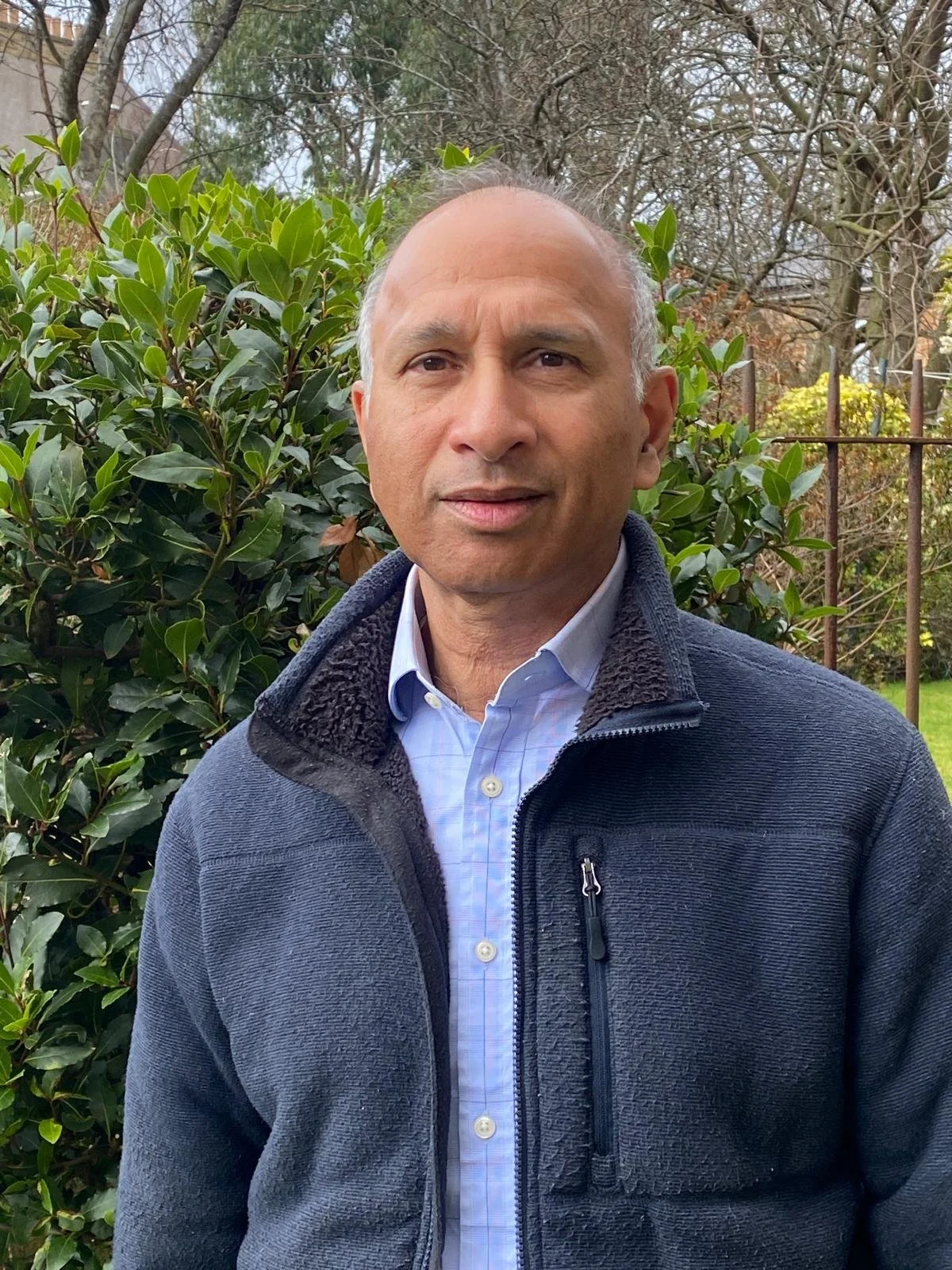 A woman with short blonde hair, wearing an orange sleeveless top and beige pants, sitting on a beige ottoman, writing in a notebook, in a well-lit room with a large leafy plant and modern abstract artwork.