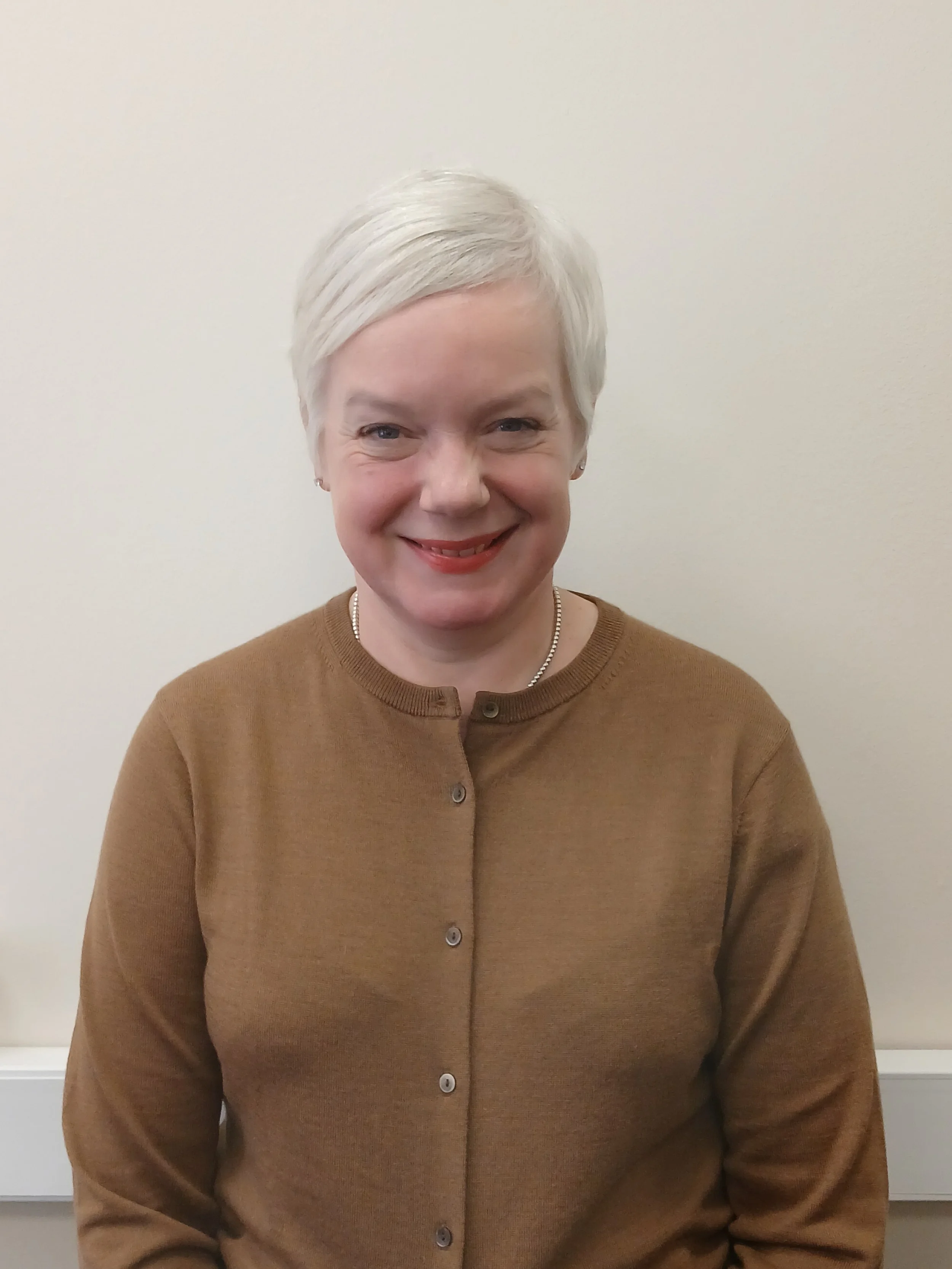 A woman with short white hair smiling in front of a plain off-white wall, wearing a light brown button-up sweater.