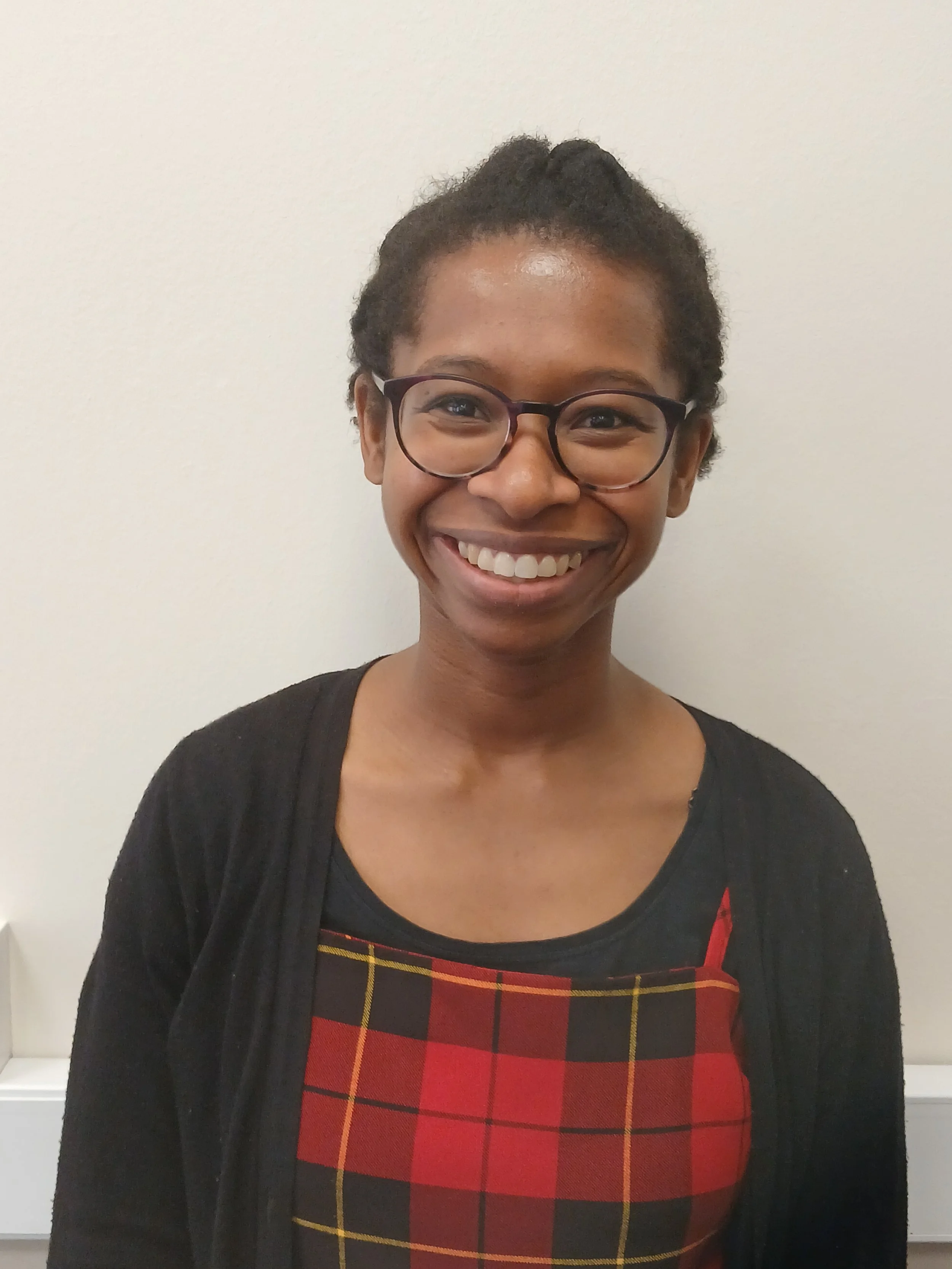 A young man with dark skin and short hair sitting on a bench against a neutral wall, wearing a gray polo shirt and cream-colored pants, with a contemplative expression and shadow cast on the wall.