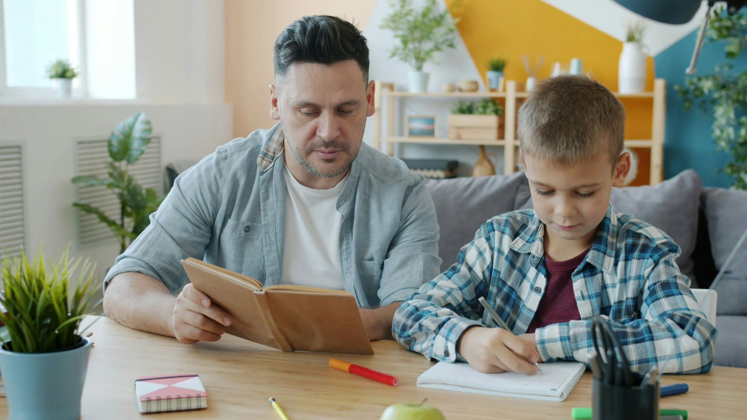 A man and a boy sitting at a wooden desk, with the man reading a book and the boy writing in a notebook, surrounded by plants and office supplies in a colourful room.
