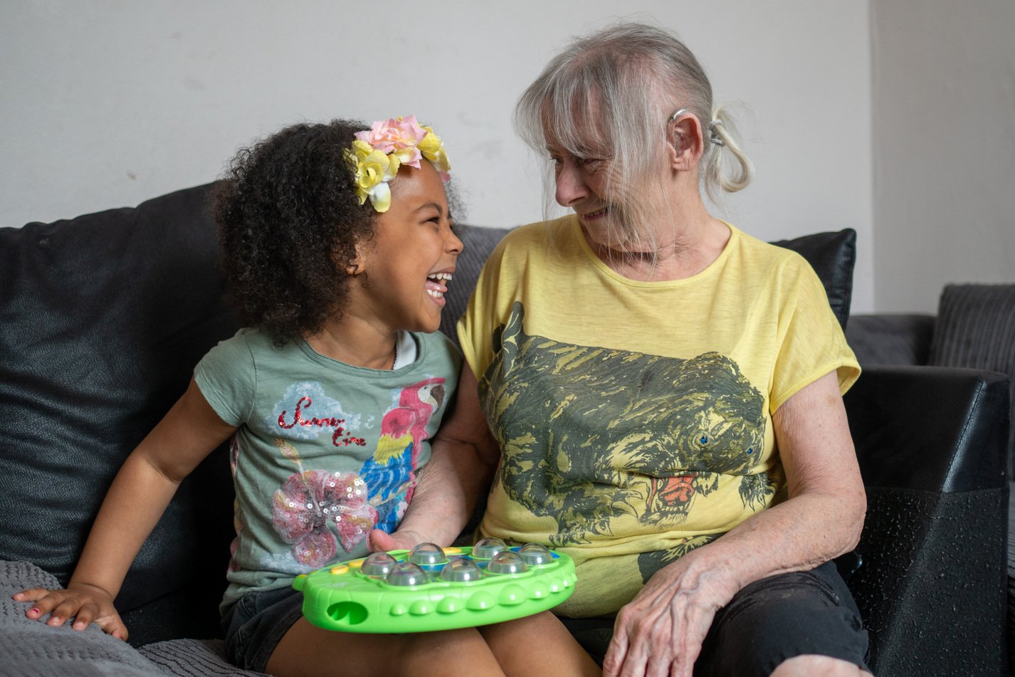 A young girl and an elderly woman sitting on a couch, smiling and enjoying each other's company, with a green toy on the girl's lap.