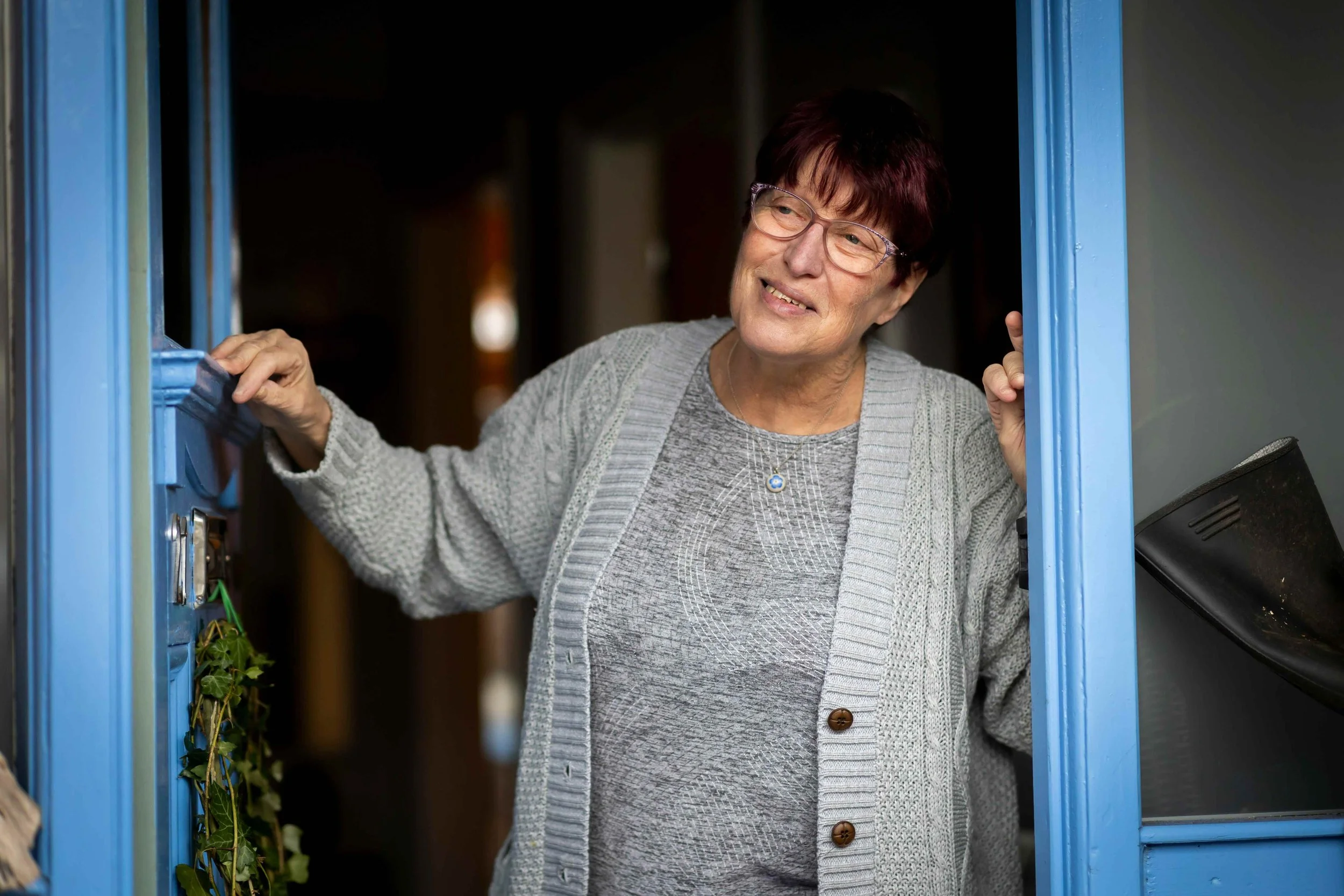 An elderly woman with short dark hair and glasses smiling while standing at her front door.