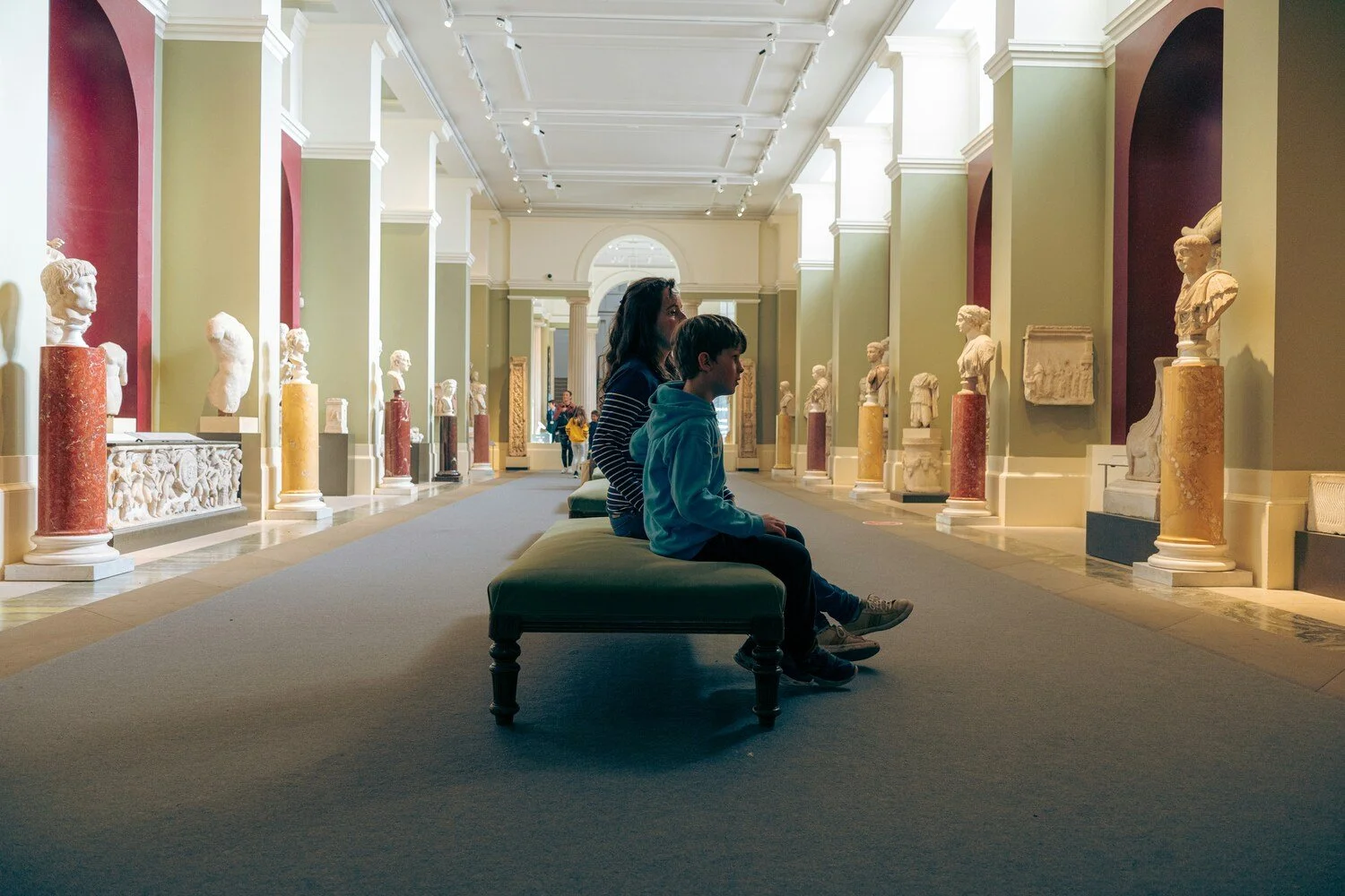A woman and a boy with dark hair sitting on a bench in an art museum, viewing ancient sculptures displayed on pedestals along the walls behind them, with other visitors visible in the background.