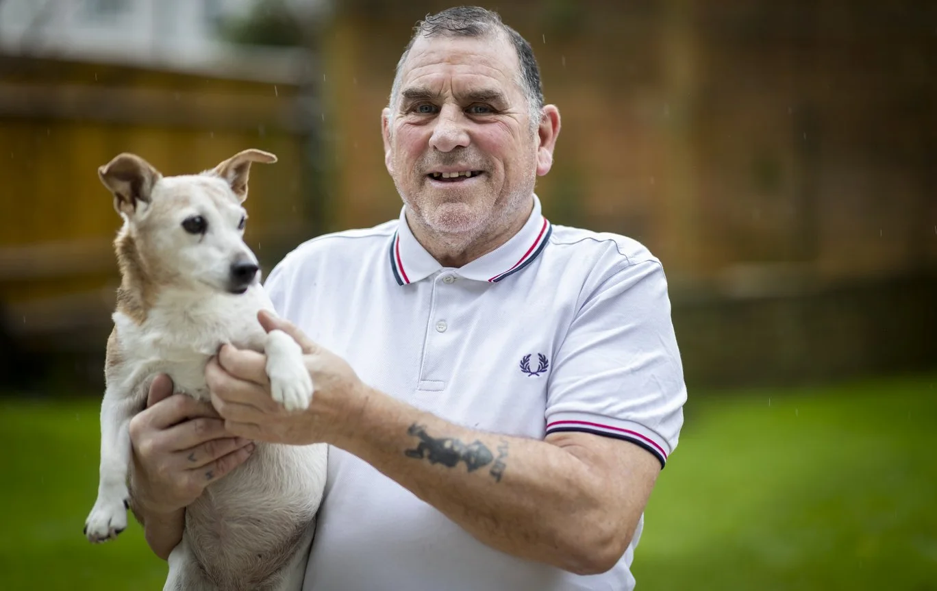 An older man with short gray hair holding a small tan and white dog outdoors in a backyard.