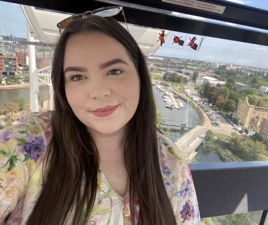 A young woman with long brown hair and a floral shirt posing outdoors with a cityscape and bridge in the background.