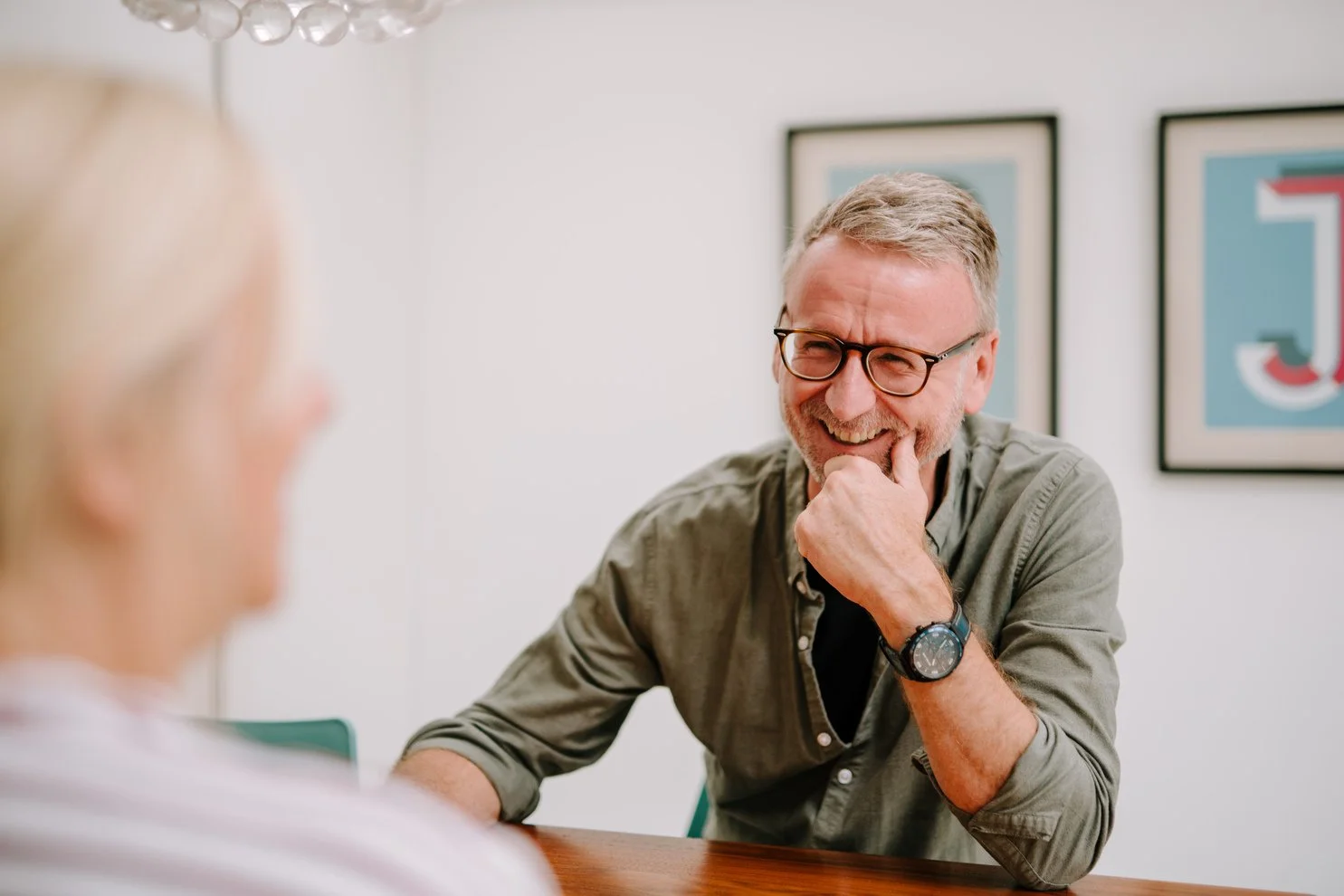 A man with gray hair, glasses, and a beard, smiling and resting his chin on his hand during a conversation with a woman whose blurred profile is visible in the foreground. The man is wearing a casual gray button-up shirt and a watch, and there are framed artwork on the wall behind him.