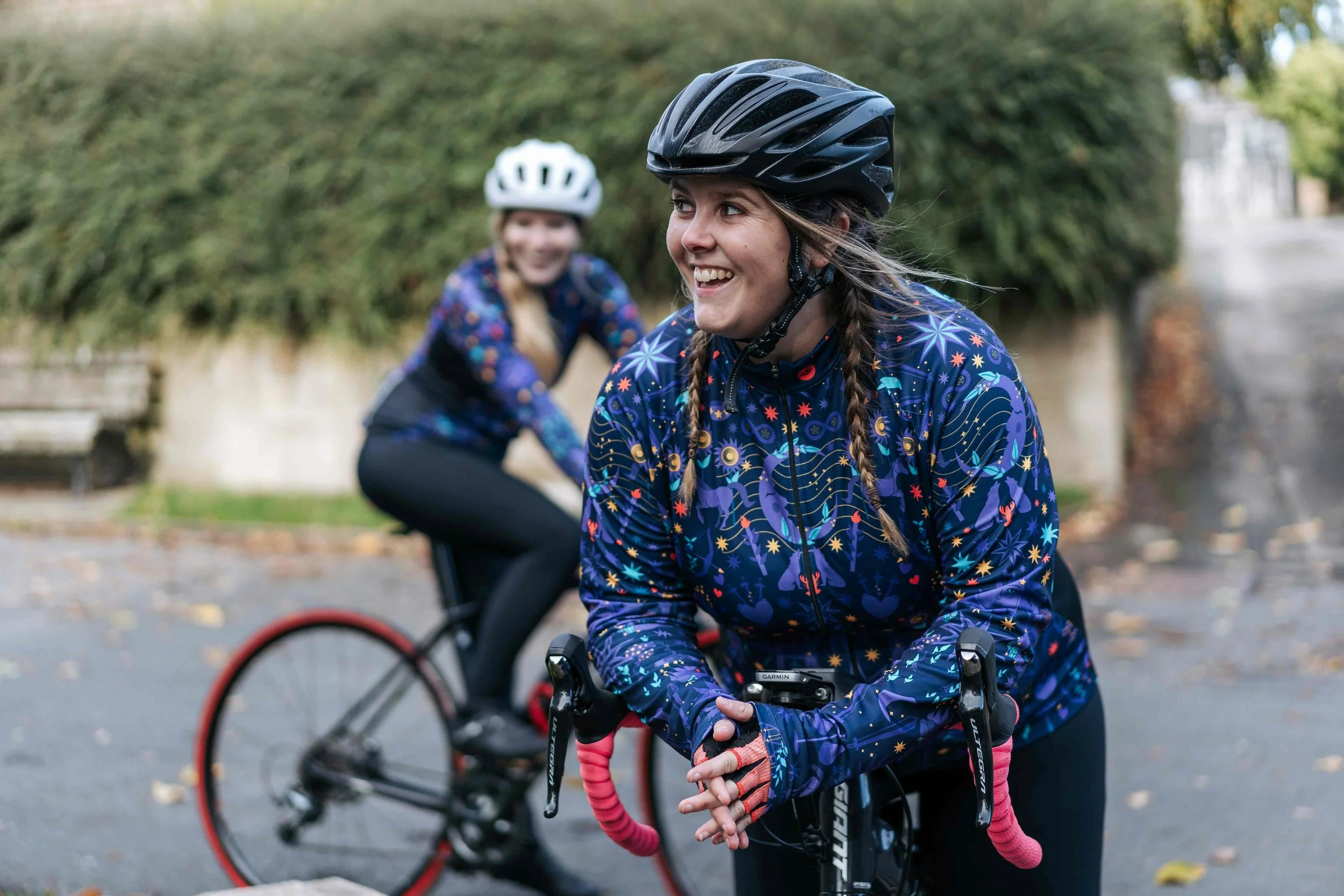 Two women in cycling gear, wearing helmets, smiling outdoors near a hedge and street, one in foreground gripping her bike's handlebar, the other in background on her bike.