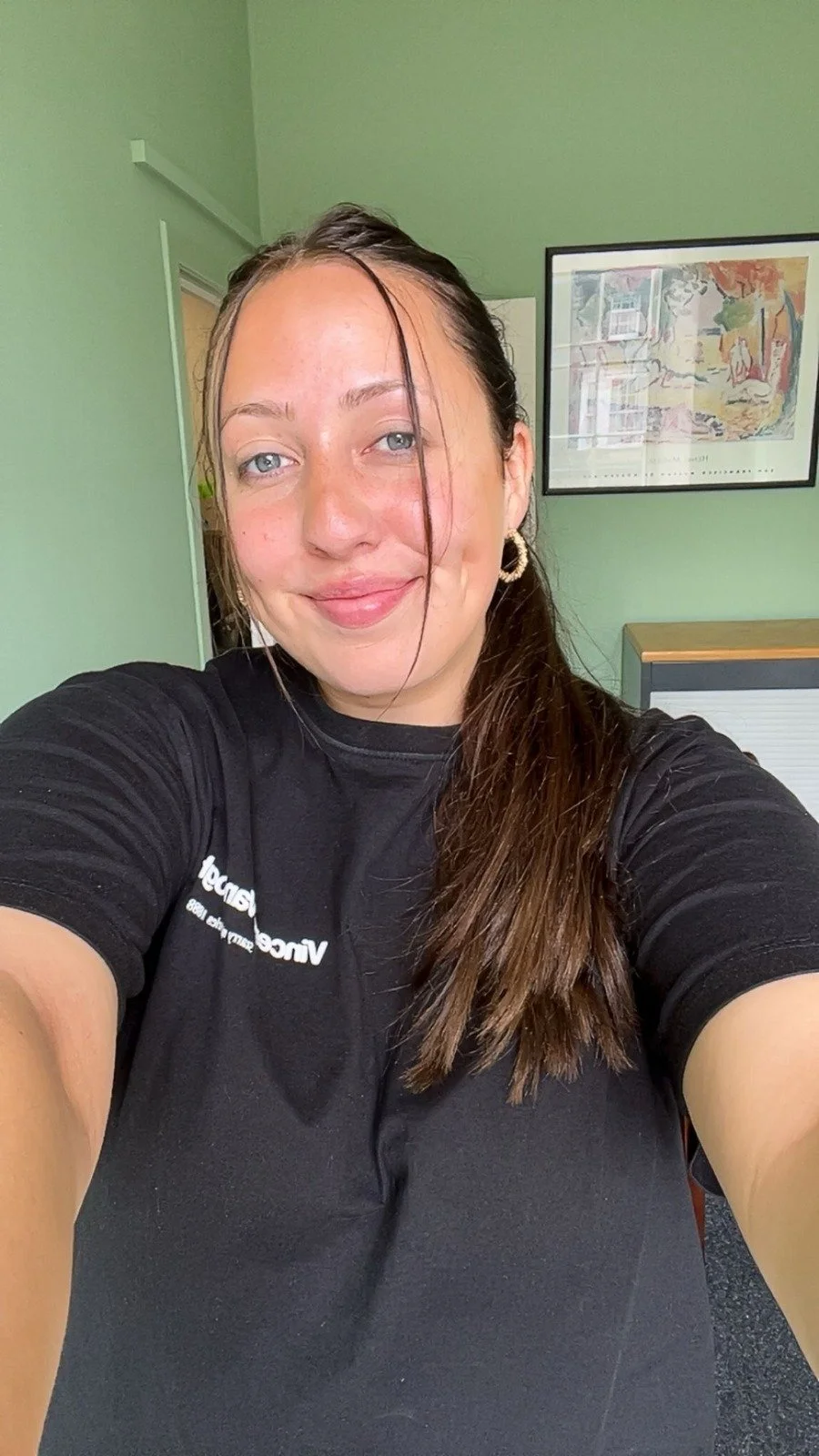 A young man with dark skin and short hair sitting on a bench against a neutral wall, wearing a gray polo shirt and cream-colored pants, with a contemplative expression and shadow cast on the wall.