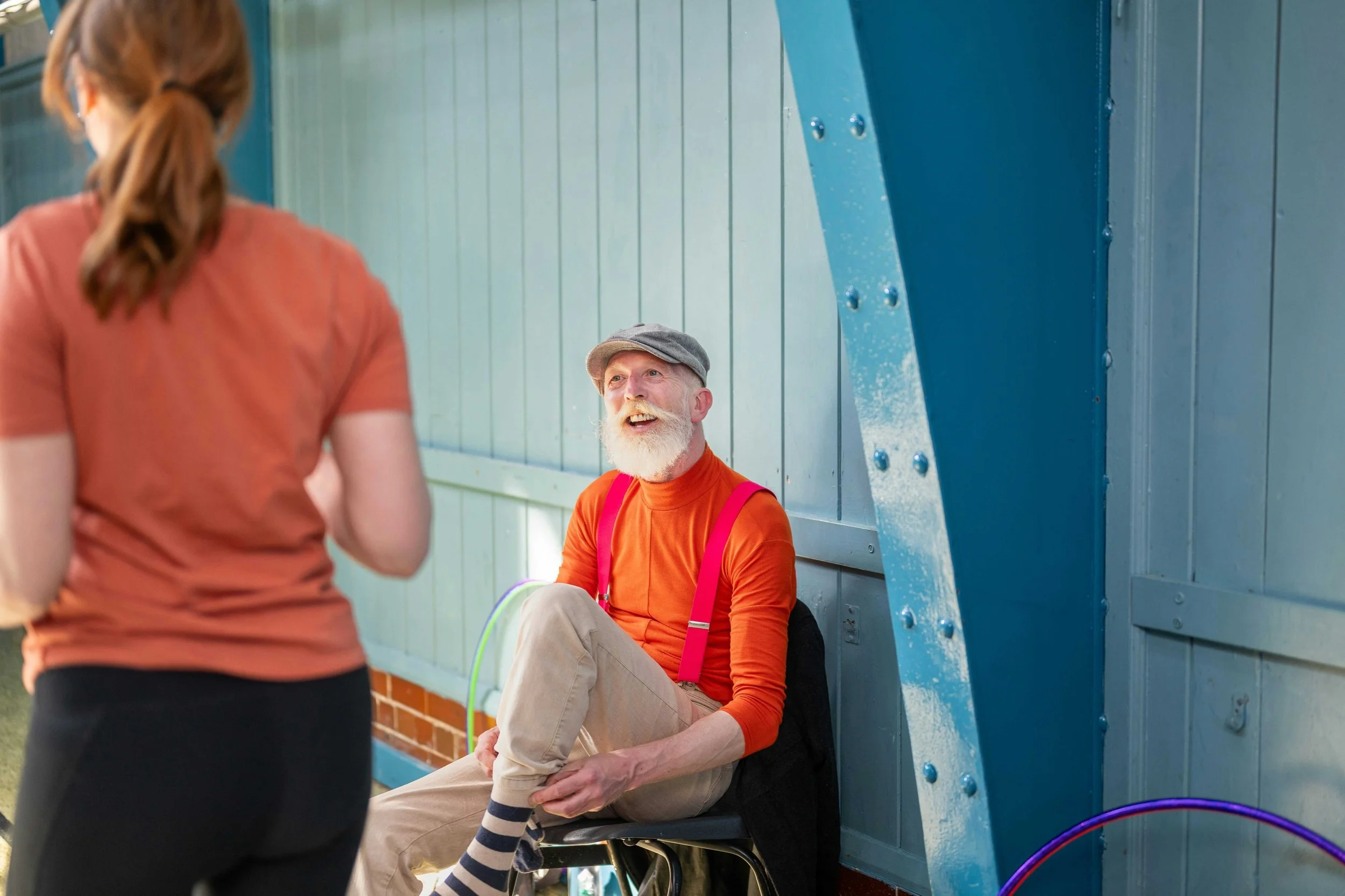 An elderly man with a white beard and gray cap sitting on a chair, talking to a woman with red hair in a ponytail wearing a rust-colored shirt, in an outdoor setting with blue walls.