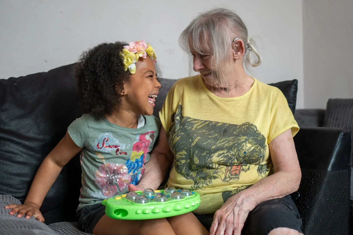 A young girl and an elderly woman sitting together on a black couch, laughing and smiling at each other. The girl is wearing a gray t-shirt with a colorful flamingo and flower design and a flower headband. She is holding a green and silver toy with clear bubbles on top. The woman is wearing a yellow t-shirt with a graphic of a roaring tiger.