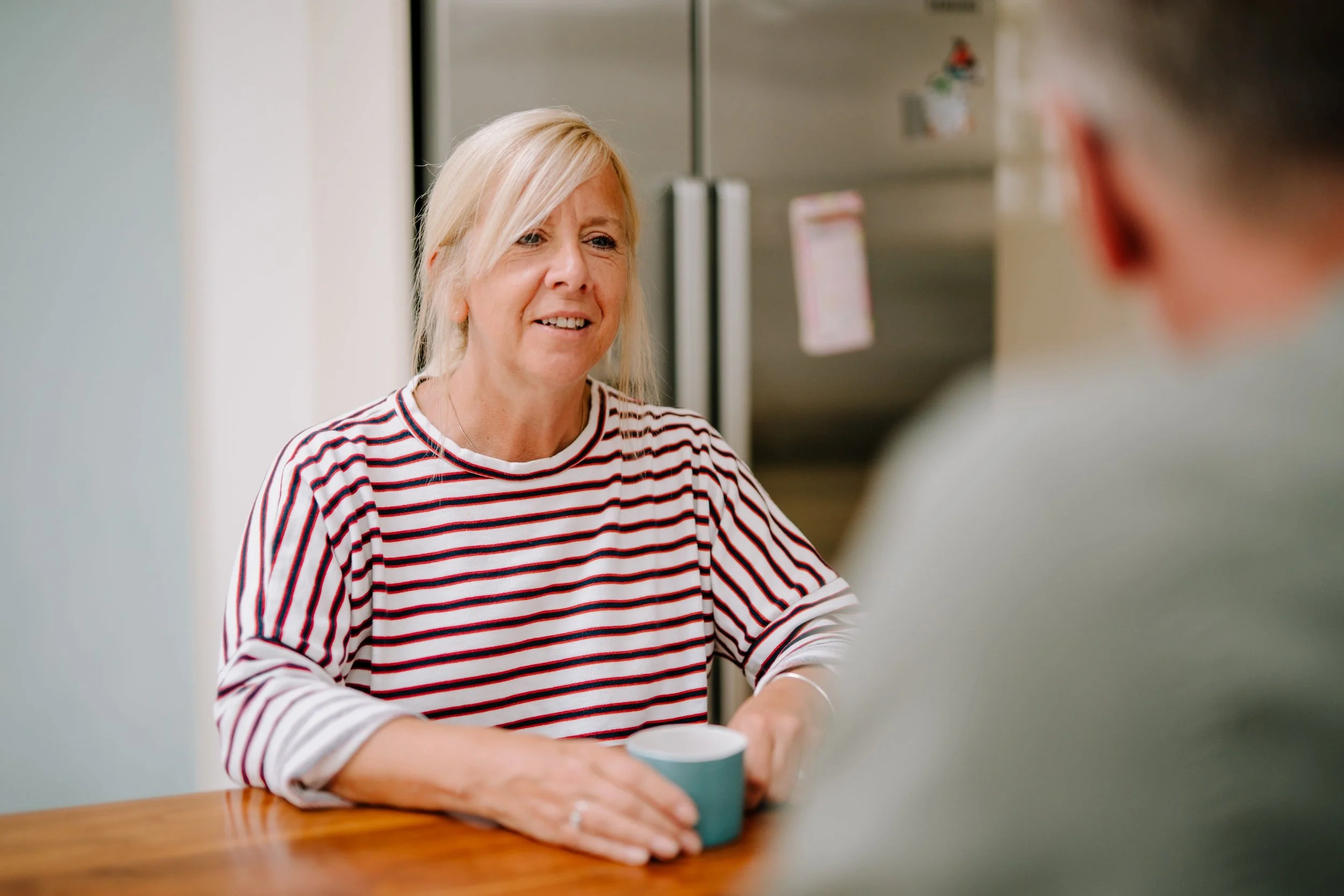 A woman with blonde hair wearing a striped shirt, holding a mug and talking to someone in a kitchen.