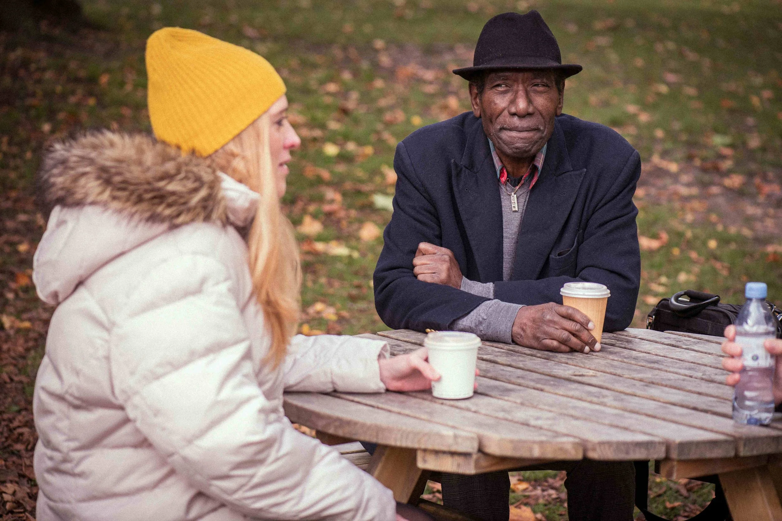 Two people sitting at a wooden picnic table outdoors, one woman and one man. The woman has long blonde hair, is wearing a yellow knit hat, a white coat with a fur-lined hood, and holding a coffee cup. The man has dark skin, is wearing a black hat, a dark jacket, and holding a coffee cup. They are in a park setting with fallen leaves.