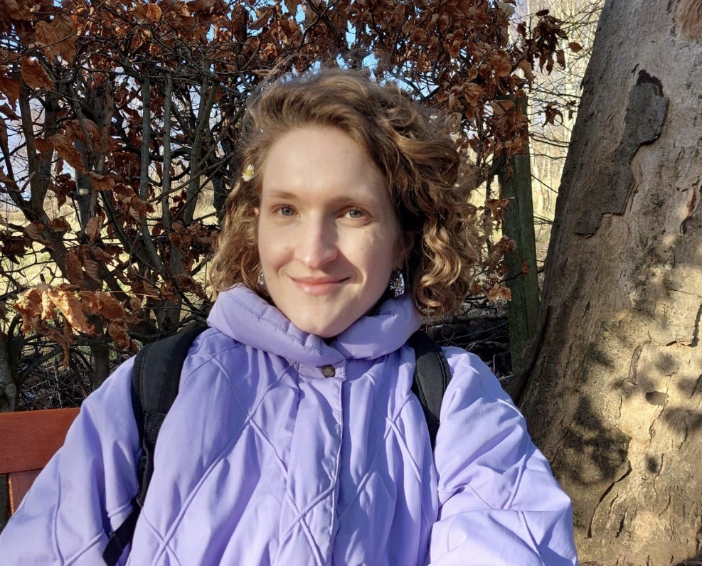 A young woman with curly brown hair smiling, wearing a black top and a gold necklace.