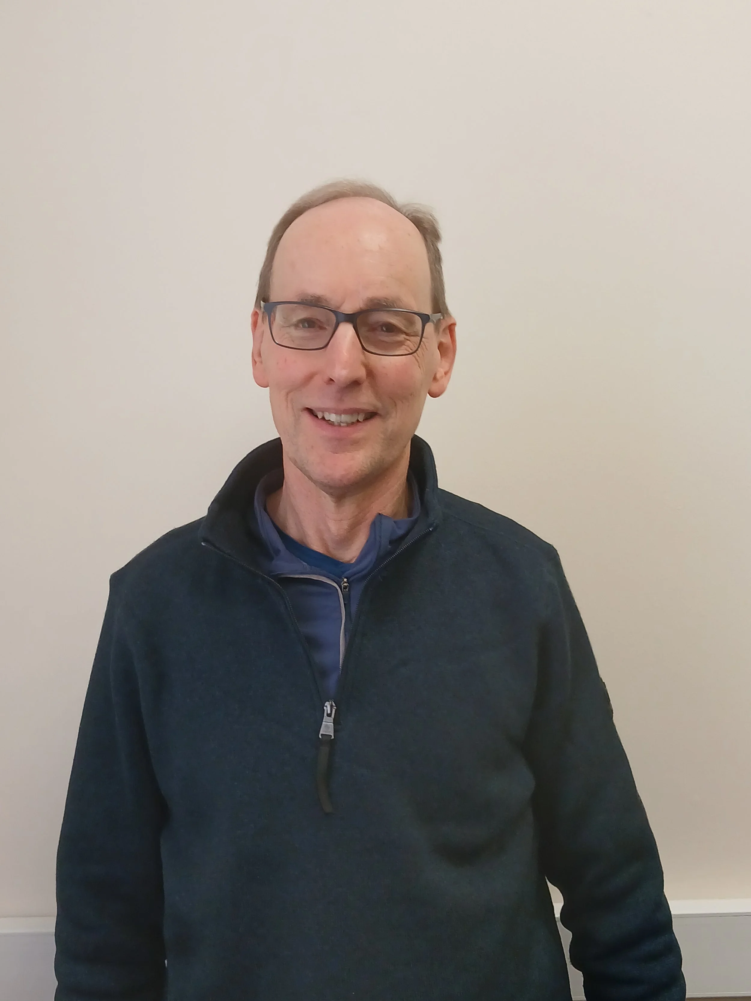 A smiling man with glasses wearing a dark blue fleece jacket standing against a plain light-colored wall.