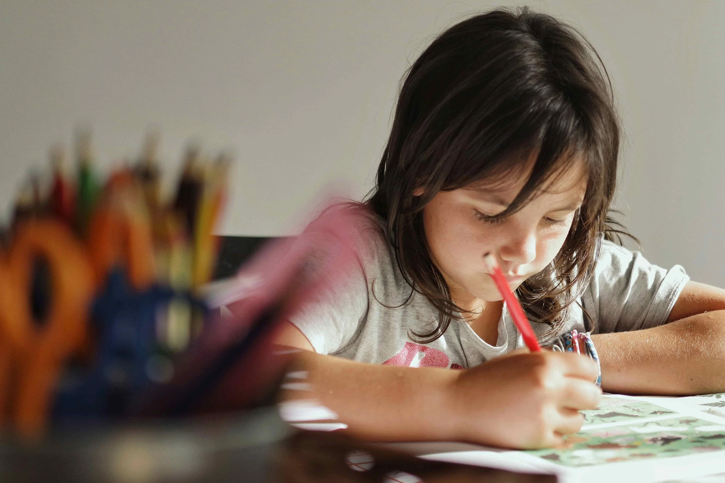 A young girl with dark hair coloring or drawing on paper with a red crayon, sitting at a table with a blurred container of colored pencils in the foreground.