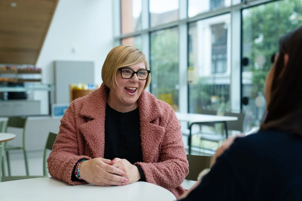 Two women sitting at a table, one with blonde hair, glasses, and a pink coat, smiling and talking.