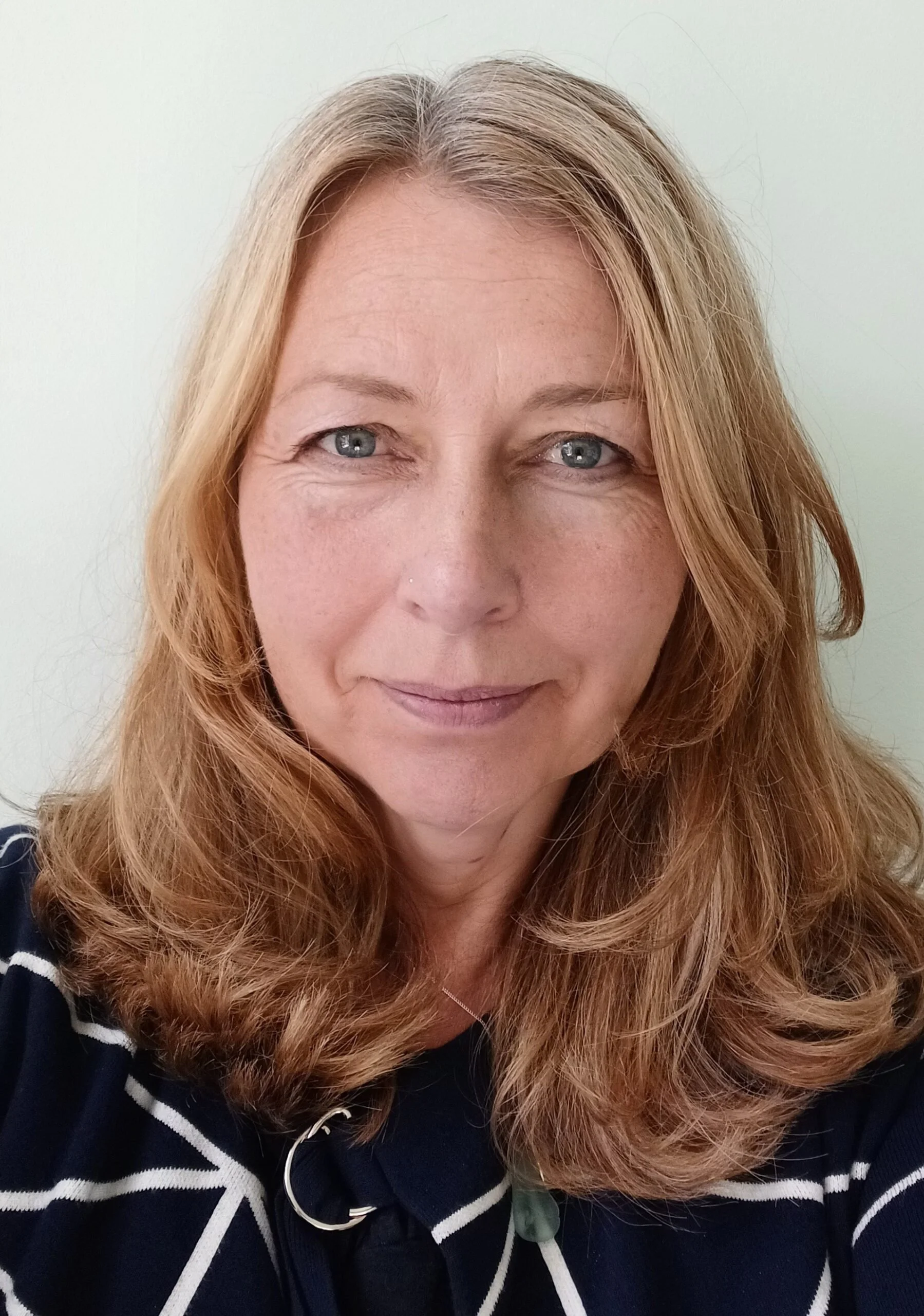 A woman with short blonde hair, wearing an orange sleeveless top and beige pants, sitting on a beige ottoman, writing in a notebook, in a well-lit room with a large leafy plant and modern abstract artwork.