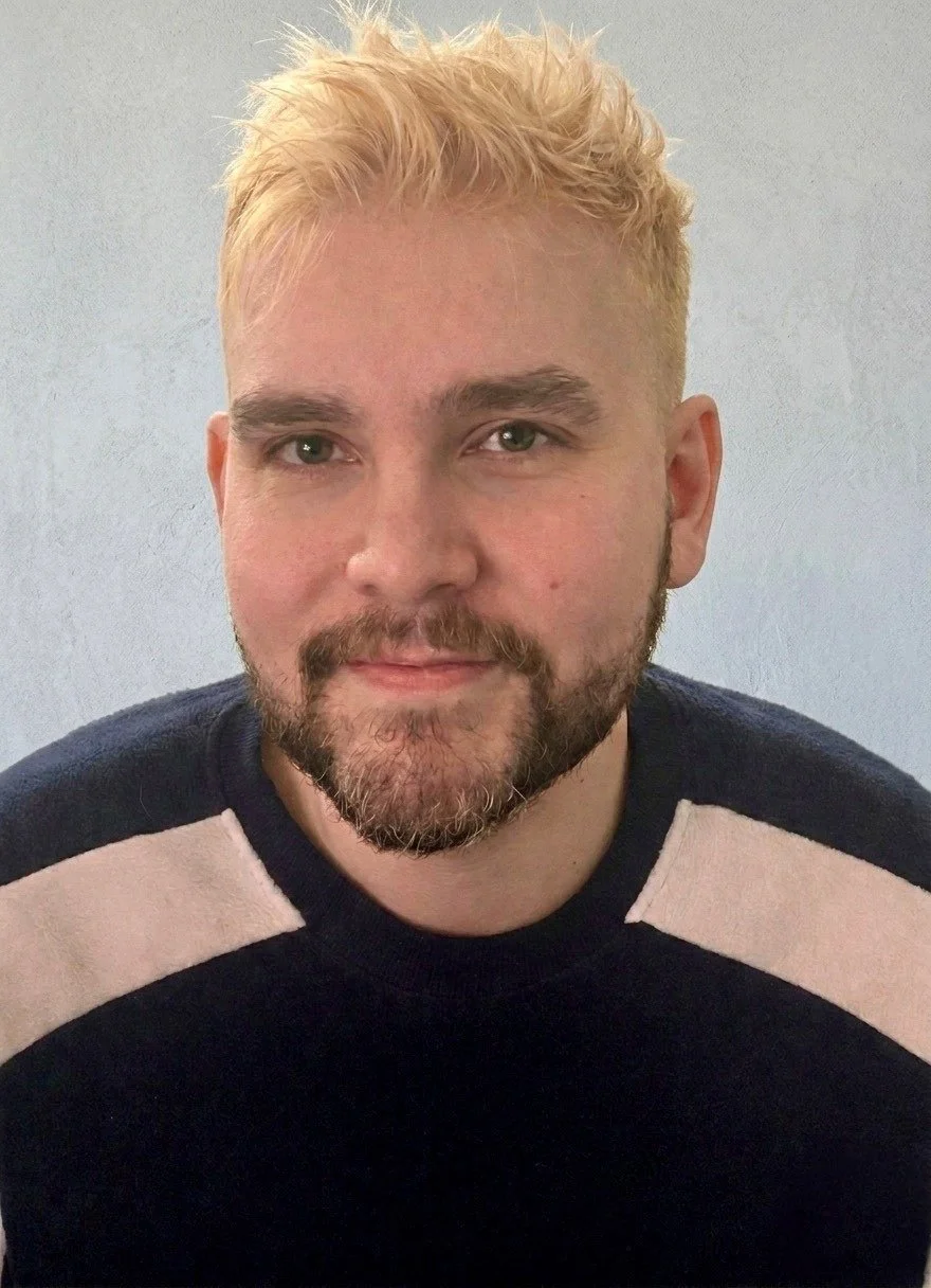 A young man with curly hair, glasses, and wearing a blue jacket and hoodie.