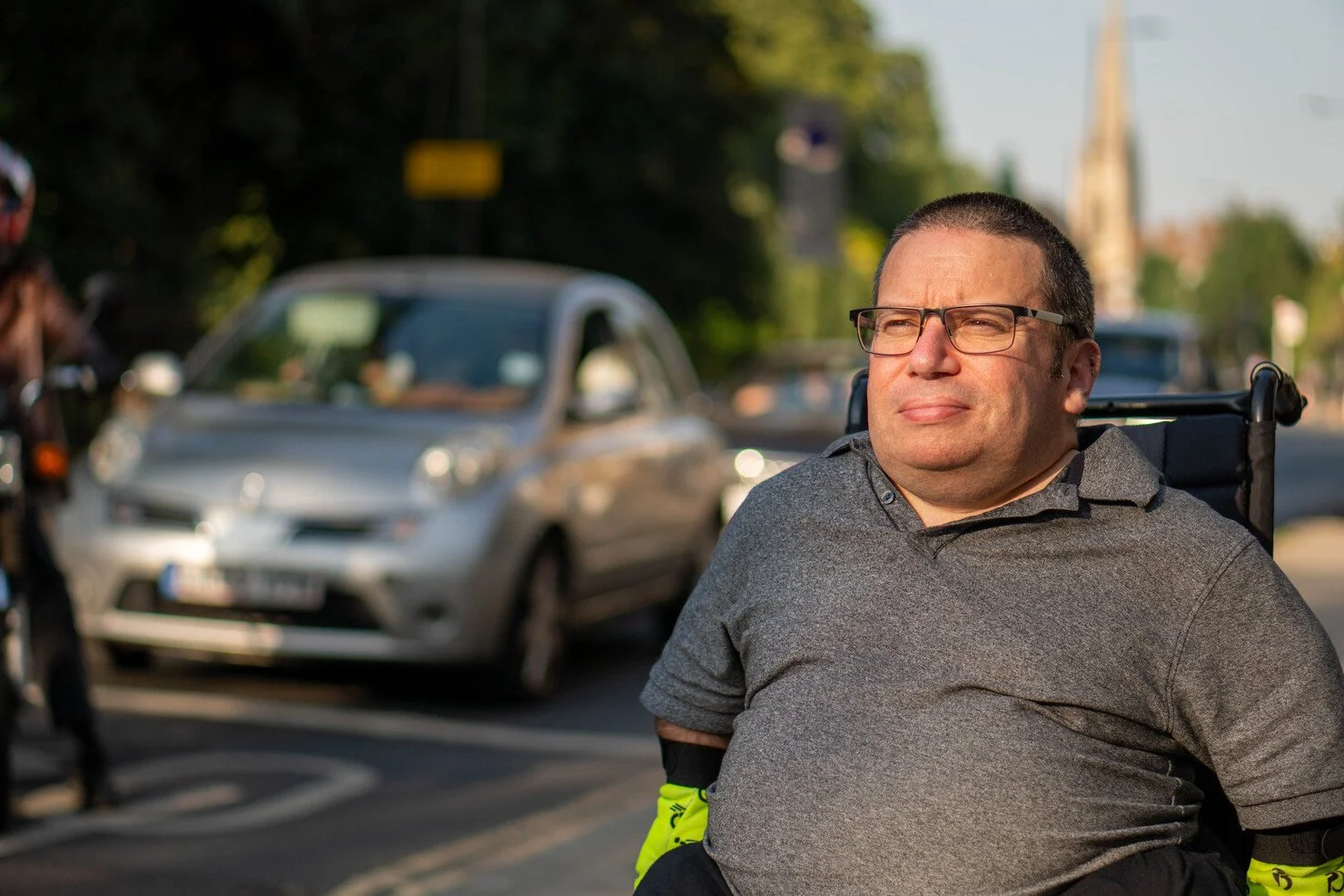 A man with glasses in a wheelchair on a road, with cars and a cyclist in the background, looking into the distance as the sun sets.