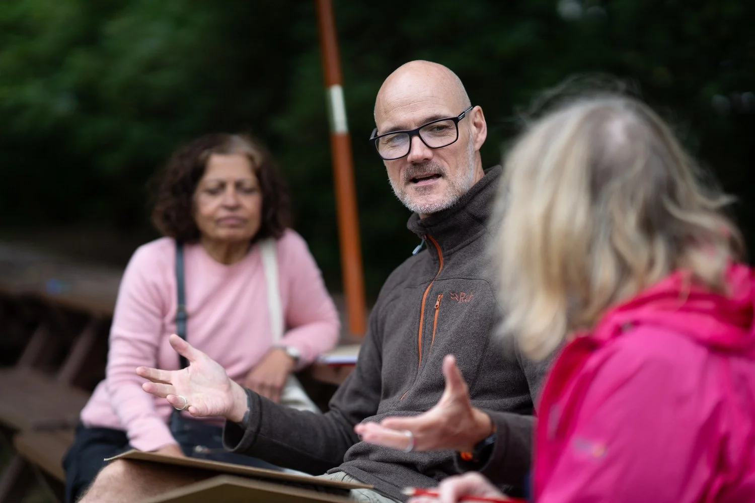 Three people sitting outdoors in conversation, with a man wearing glasses and two women, one with curly hair and the other with blonde hair, engaged in discussion.