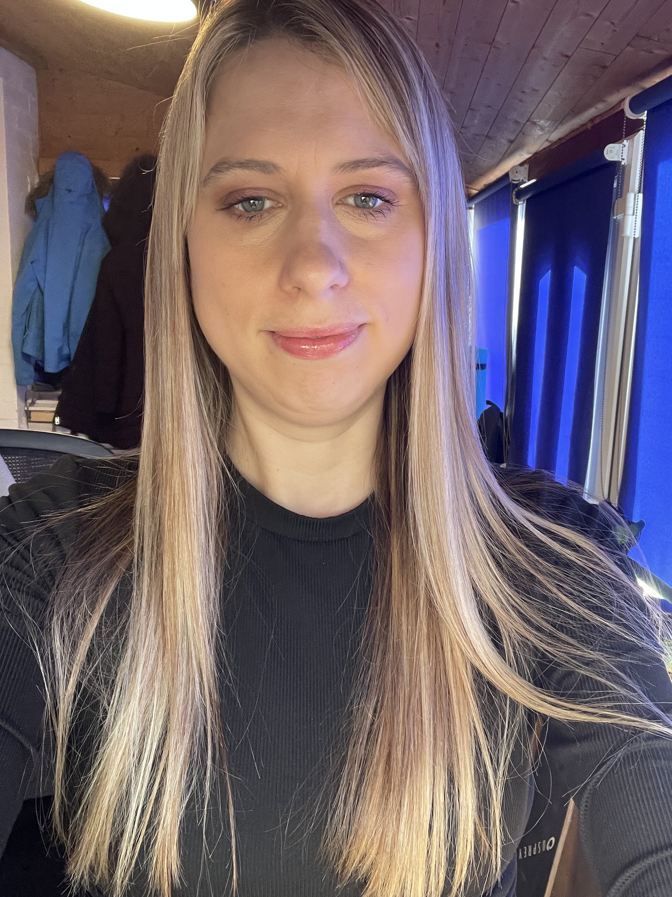 A woman with long blonde hair wearing a black top, taking a selfie in a room with blue window blinds and a wooden ceiling.