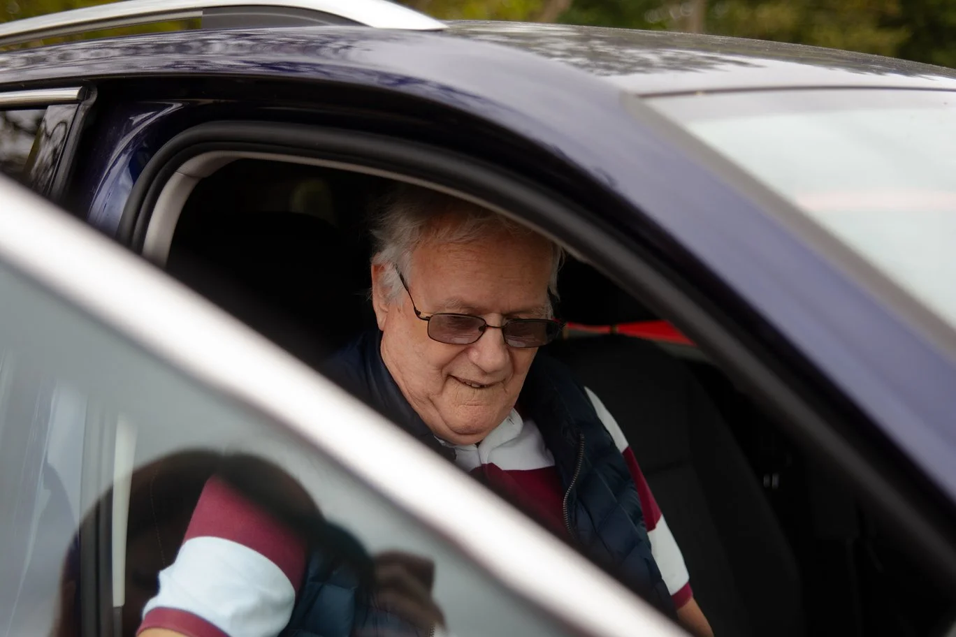 An elderly man with glasses looks down while sitting in the driver's seat of a car.