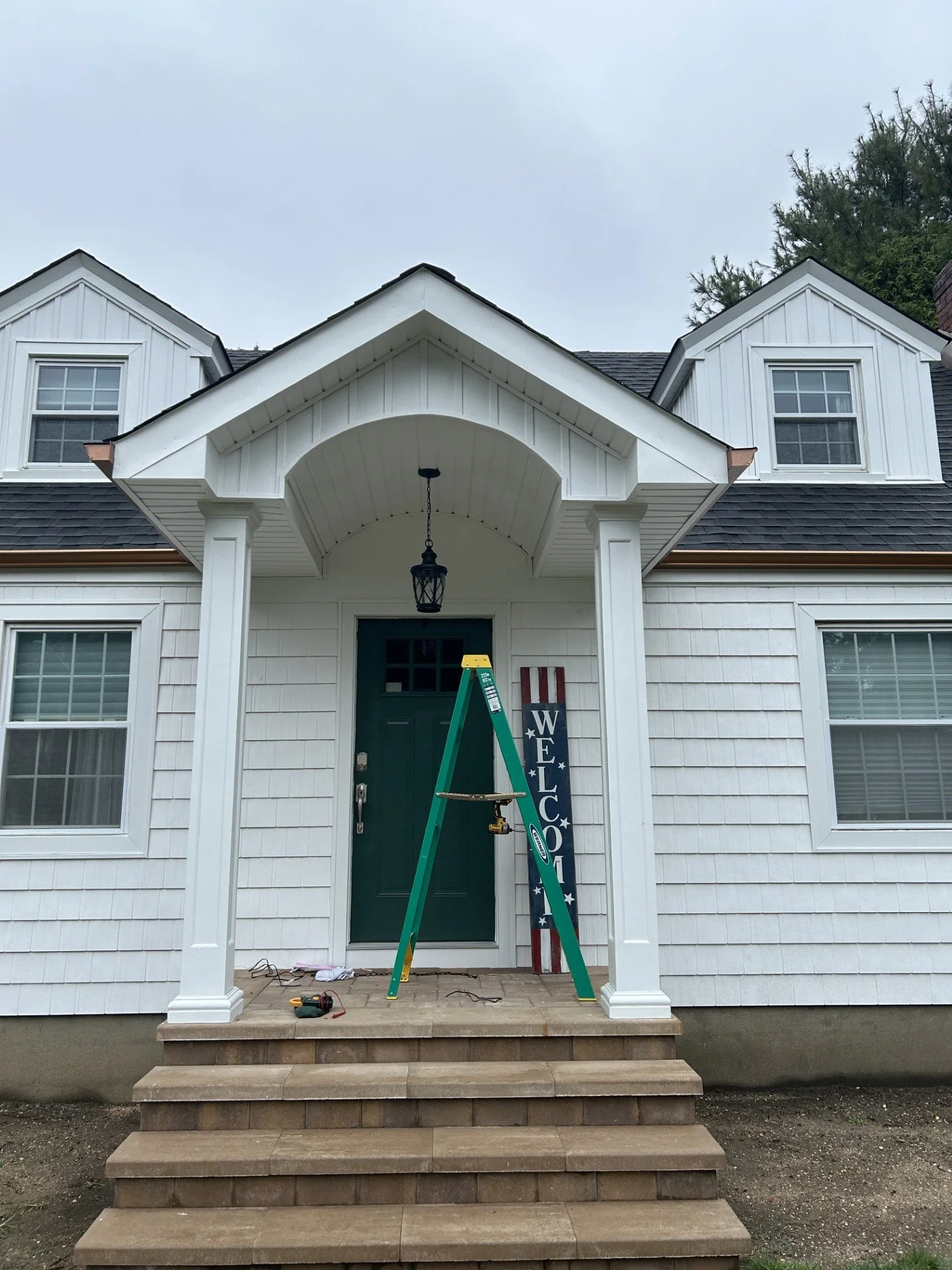 House with white exterior, green front door, steps leading to porch, ladder and welcome sign near the entrance, overcast sky.