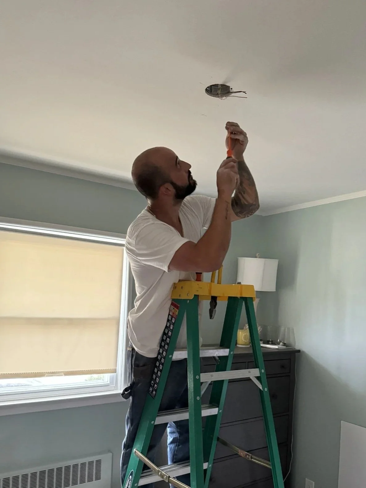 A man standing on a step ladder working on a ceiling light fixture in a room with gray walls and a window with beige blinds.