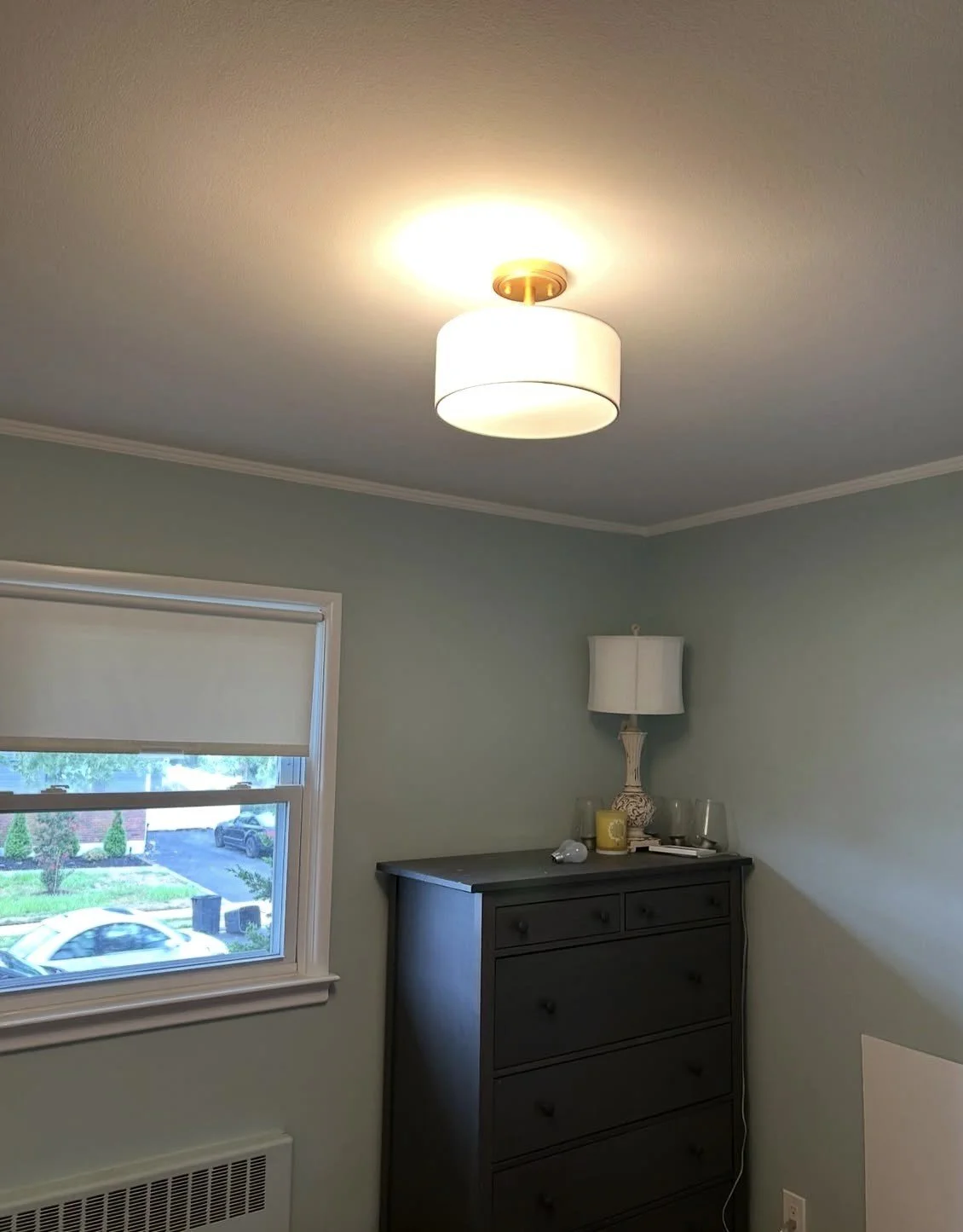 A room with a ceiling light, a window with a white roller shade, a black dresser, and a table lamp on top of the dresser.