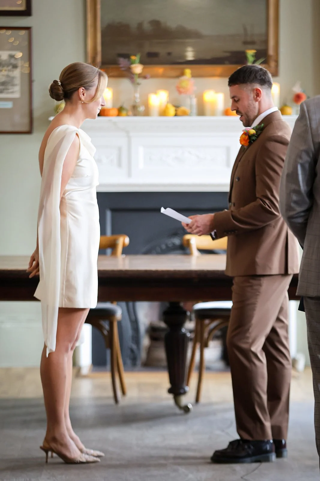 A woman in an elegant white dress and clear heels stands in front of a man in a brown suit, holding a paper, during a wedding ceremony in a decorated indoor venue.