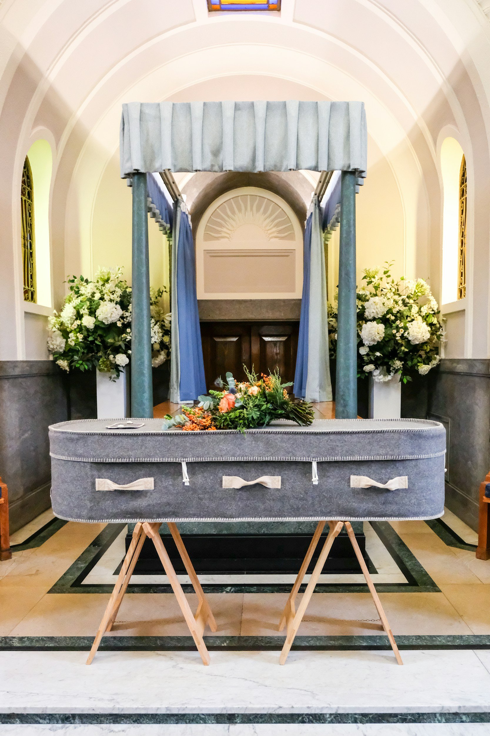 A wedding setup in a church with a floral arrangement on the altar, blue drapes forming an arch, and a gray prayer bench with wooden support stands in the foreground.