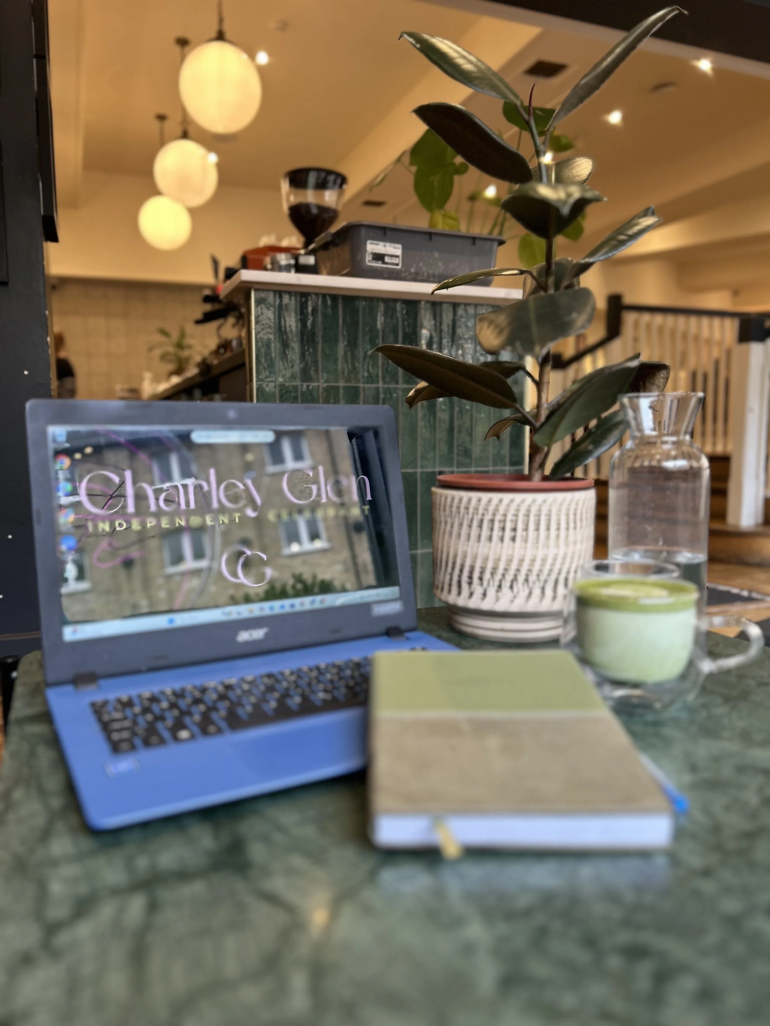 A laptop showing a website for Charlie's Glen, a potted plant, a glass jar with water, a small cup of green tea, and a notebook on a green marble table in a cozy cafe or restaurant.