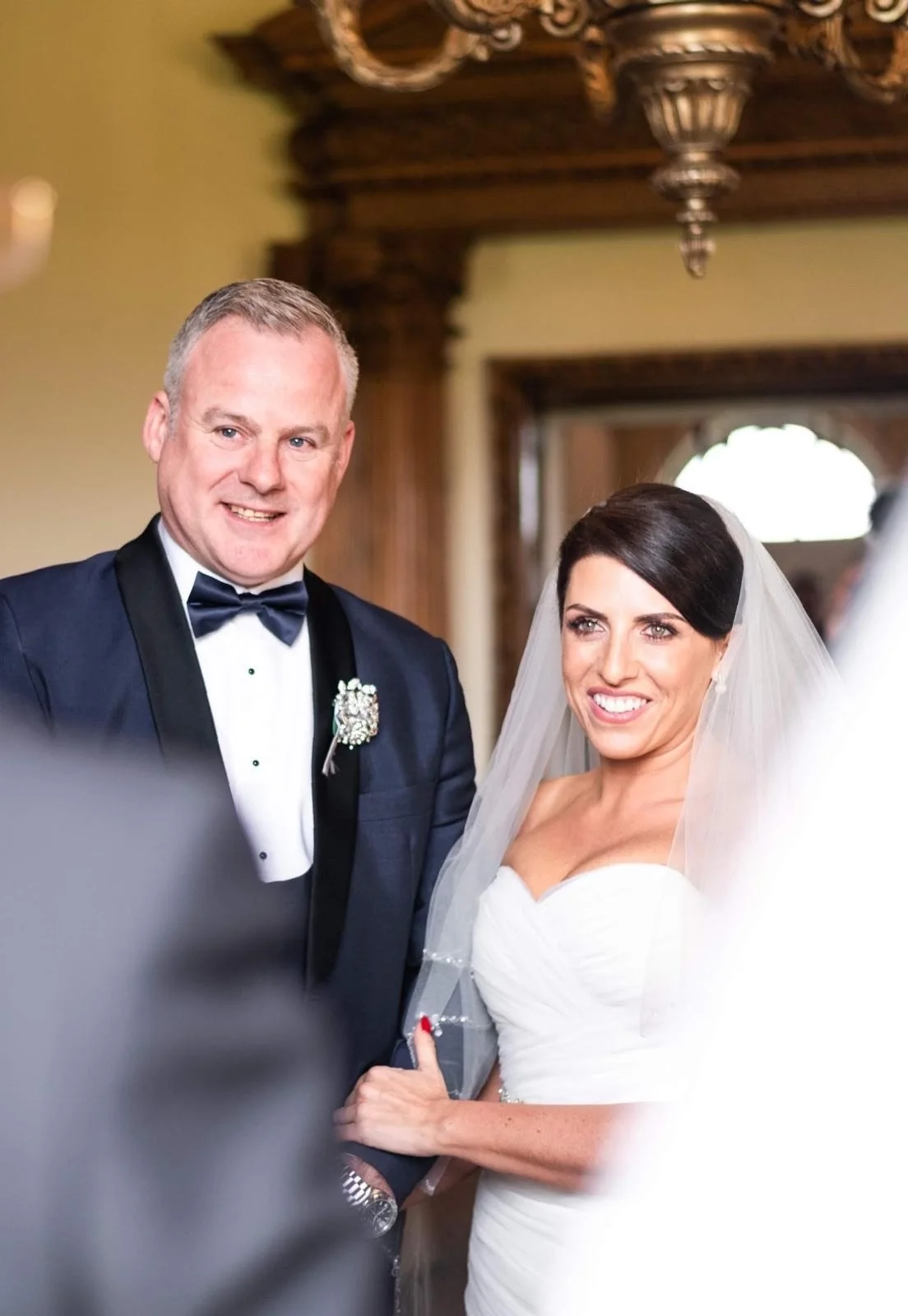 A bride and groom smiling during their wedding ceremony, standing close together indoors with ornate woodwork in the background.