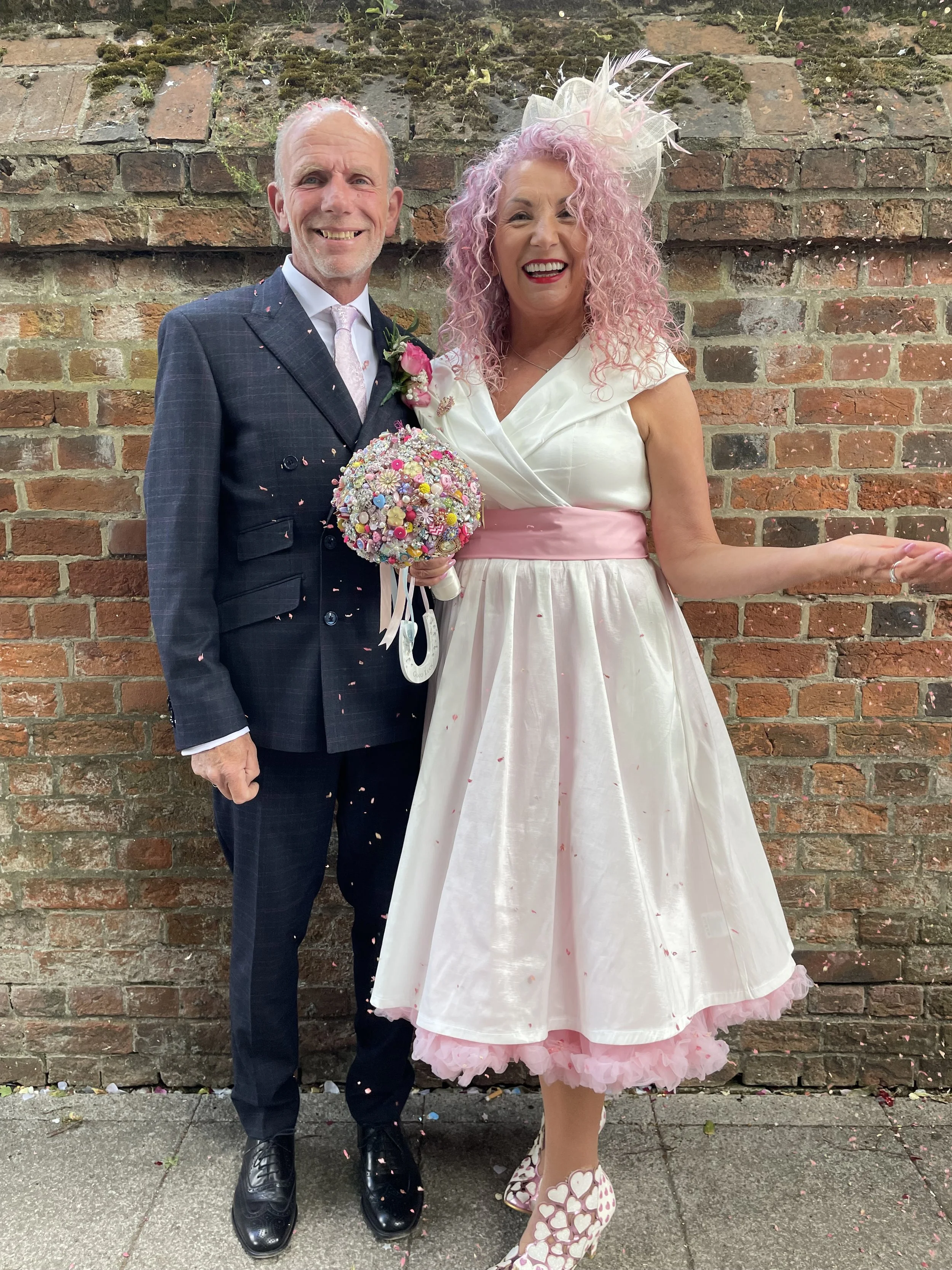 A smiling bride with pink curly hair in a white and pink dress holding a colorful bouquet, standing next to a smiling groom in a dark suit, against a brick wall background. Confetti is falling around them.