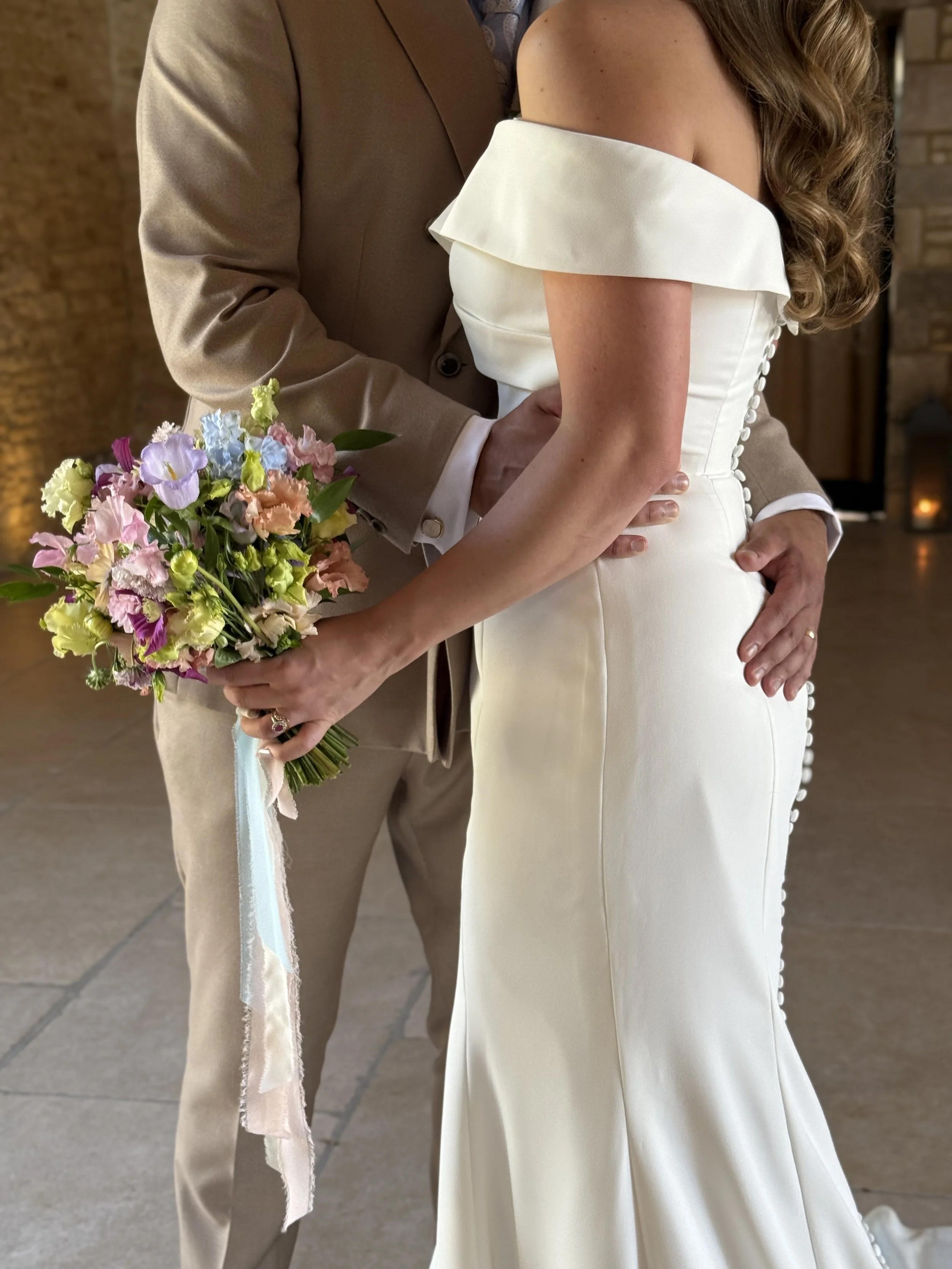 Close-up of a bride and groom holding hands during their wedding, with the bride holding a bouquet of colorful flowers. The bride is wearing an off-the-shoulder white wedding dress with buttons down the back, and the groom is dressed in a beige suit.