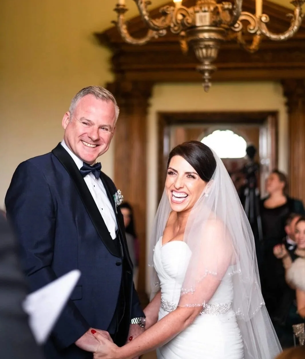 A bride and groom holding hands during their wedding ceremony, smiling and laughing, with guests in the background and an ornate chandelier overhead.