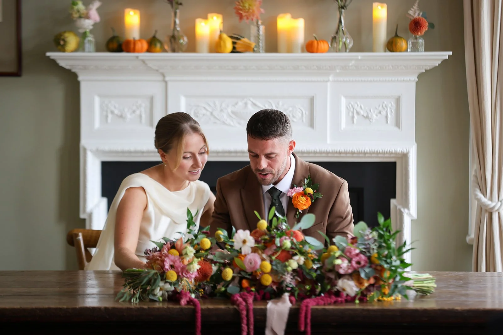 A bride and groom signing a wedding register at a table decorated with flowers, in front of a fireplace mantel with candles and pumpkins, in a cozy, decorated room.