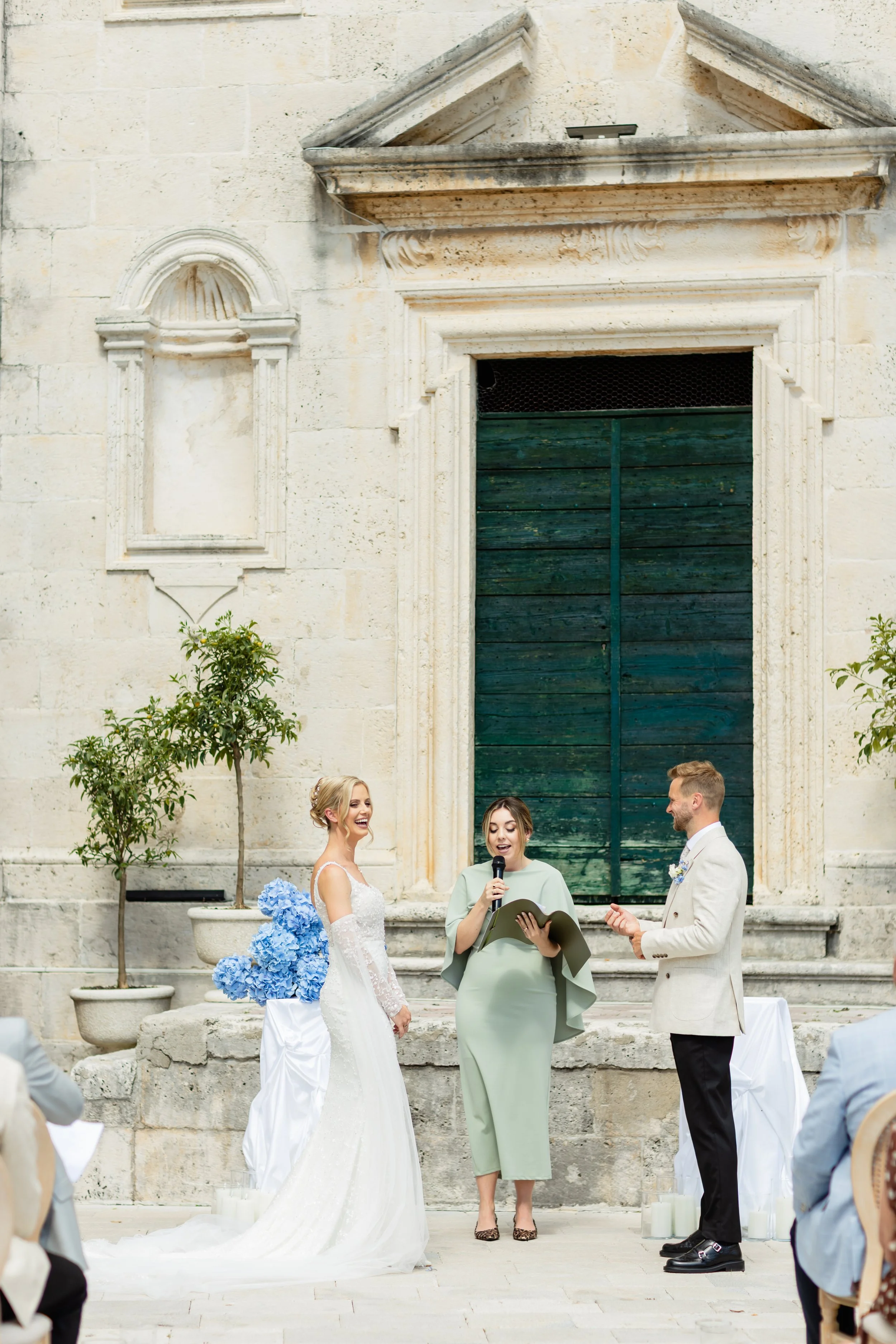 A wedding ceremony outdoors in front of an old stone building with a green door, featuring a bride in a white wedding dress and lace gloves, a groom in a light-colored suit, and an officiant woman holding a microphone and reading from a book. There are potted plants and candles around them.