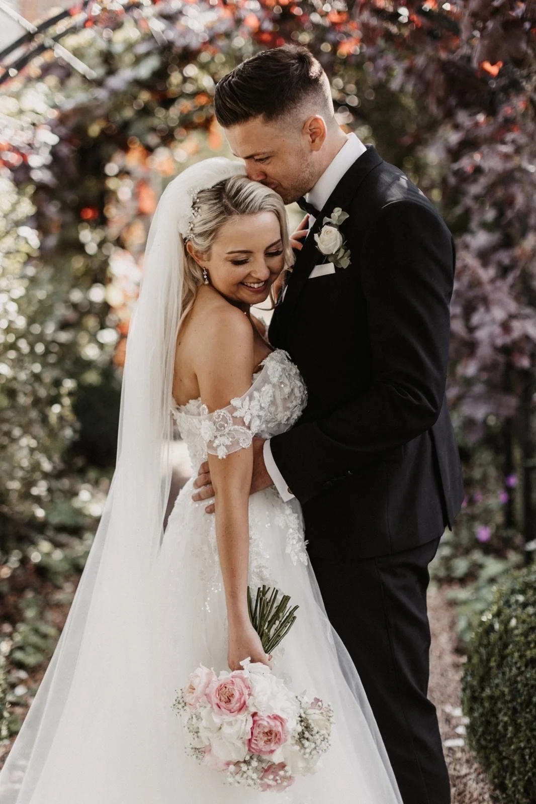 A newlywed couple smiling and embracing outdoors, with the bride holding a bouquet of pink and white roses, and the groom kissing her forehead in a garden setting with colorful trees.