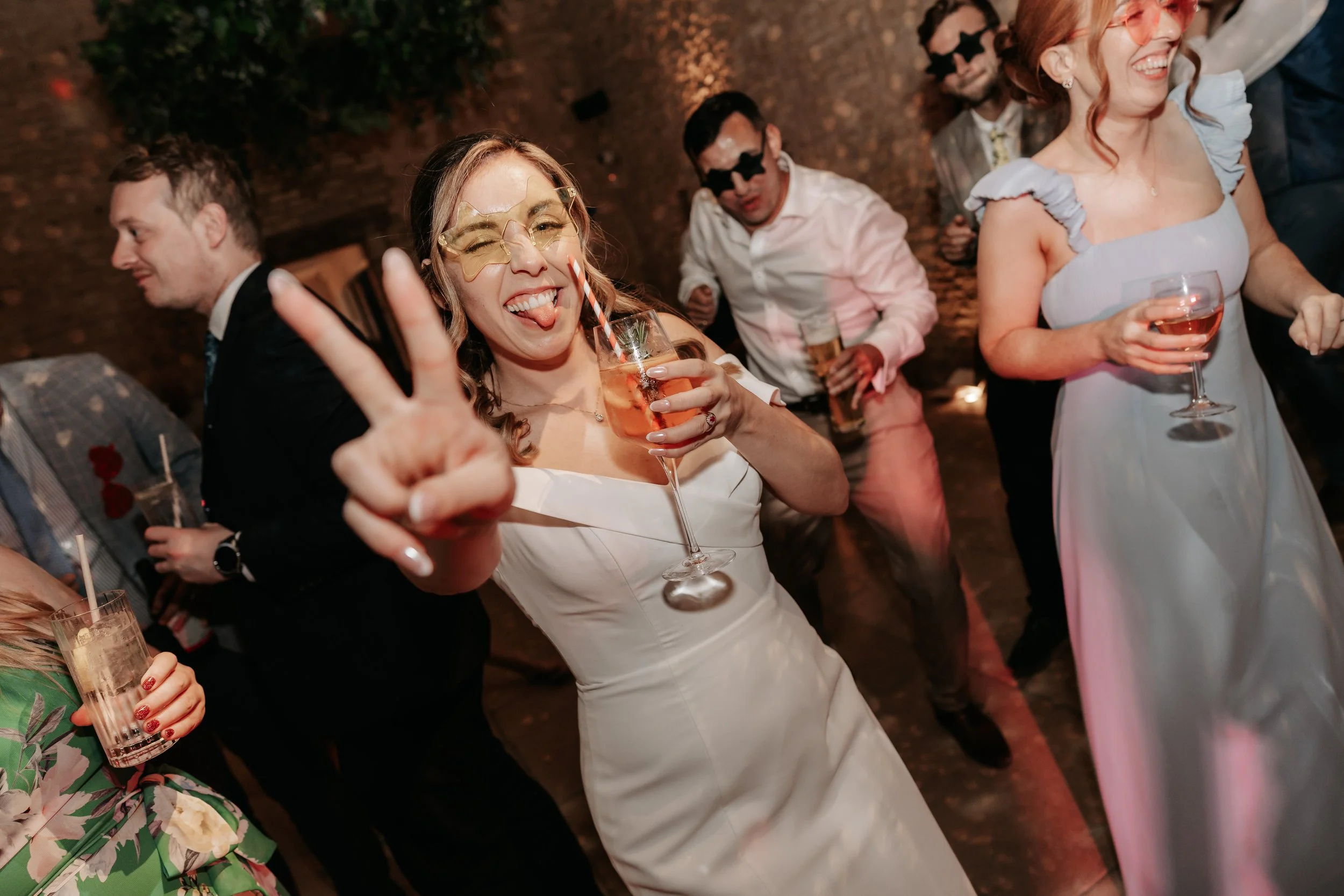 A group of people celebrating at a party. One woman in the center is wearing a white dress, gold sunglasses, and sticking out her tongue, making a peace sign with her hand while holding a glass of drink. Other guests are smiling and holding drinks, some wearing sunglasses, in a party setting with warm lighting.