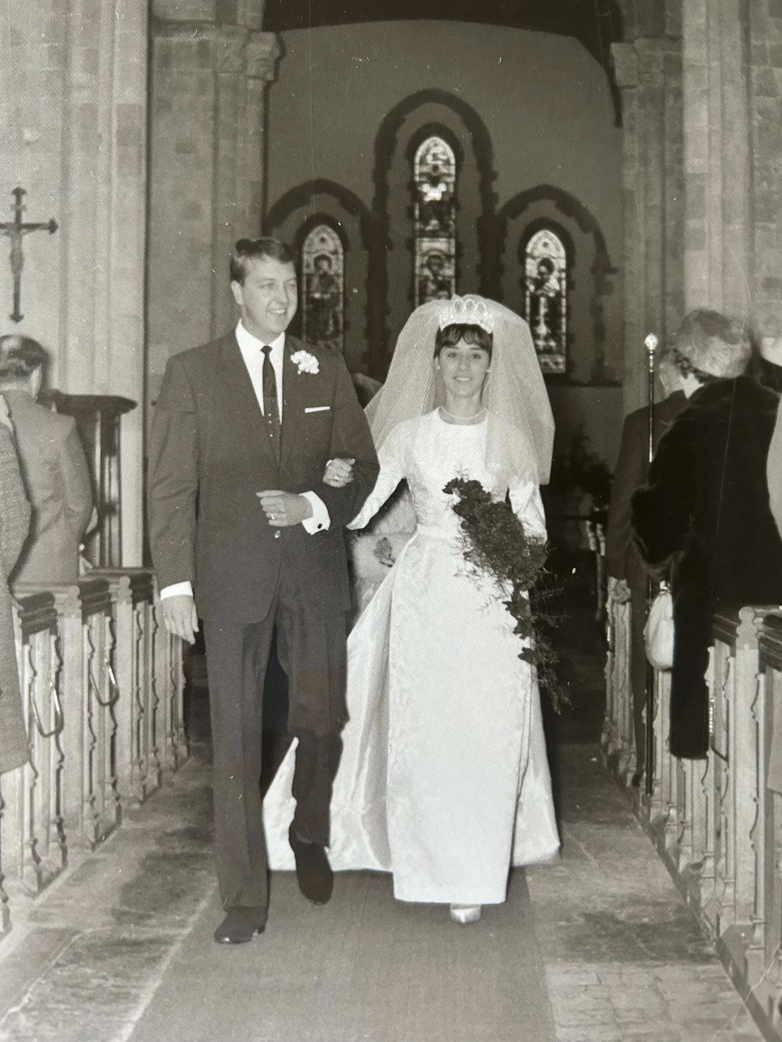 Black and white photograph of a bride and groom walking down the aisle in a church, with the bride wearing a wedding dress and veil, and the groom in a suit. They are holding hands and smiling.