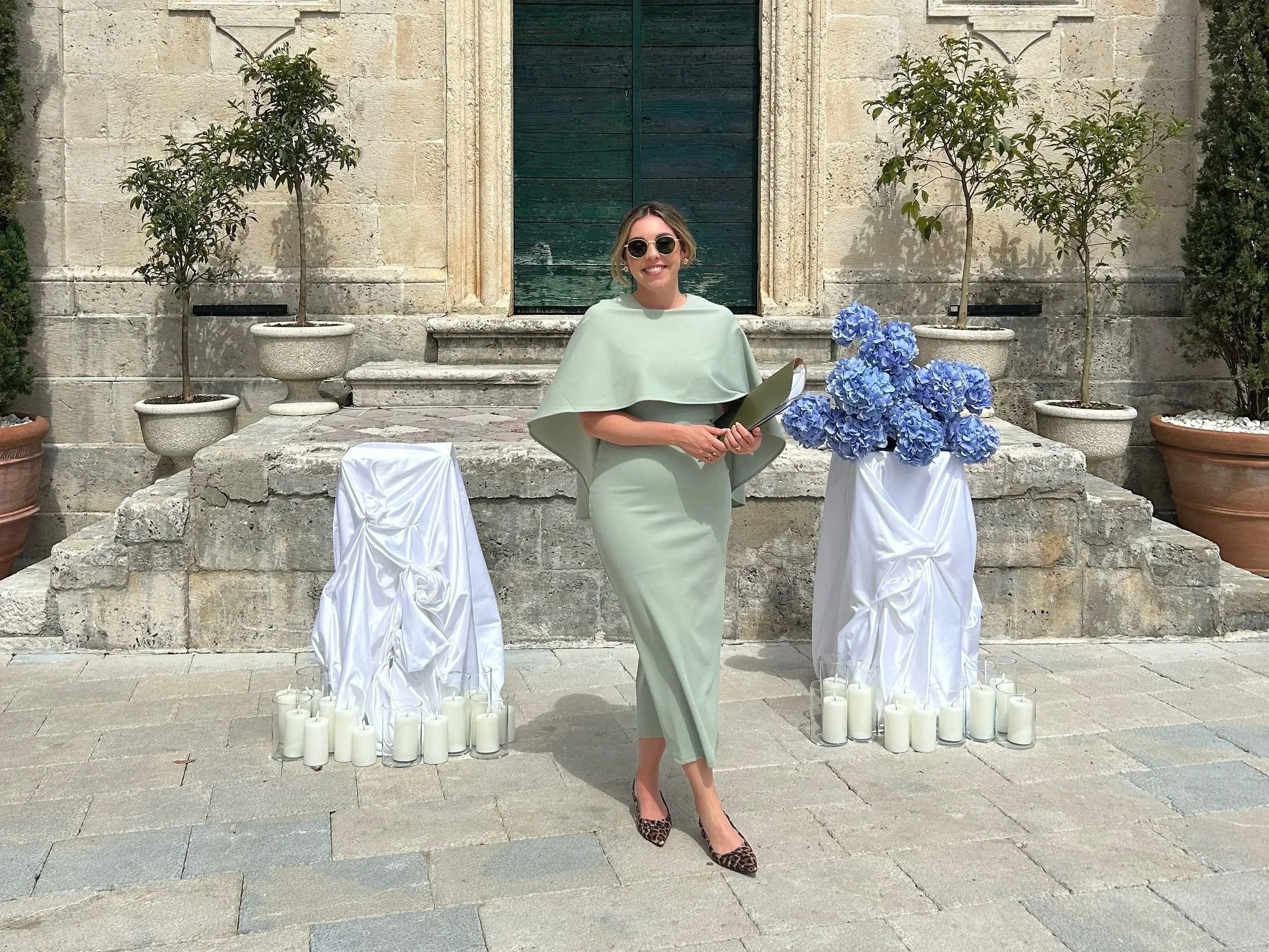 A woman in a light green dress and leopard-print shoes stands in front of a stone building with a green door. She is smiling and holding a folder or notebook. Around her are white draped chairs and numerous white candles on the ground. There are four potted plants with green foliage behind her.