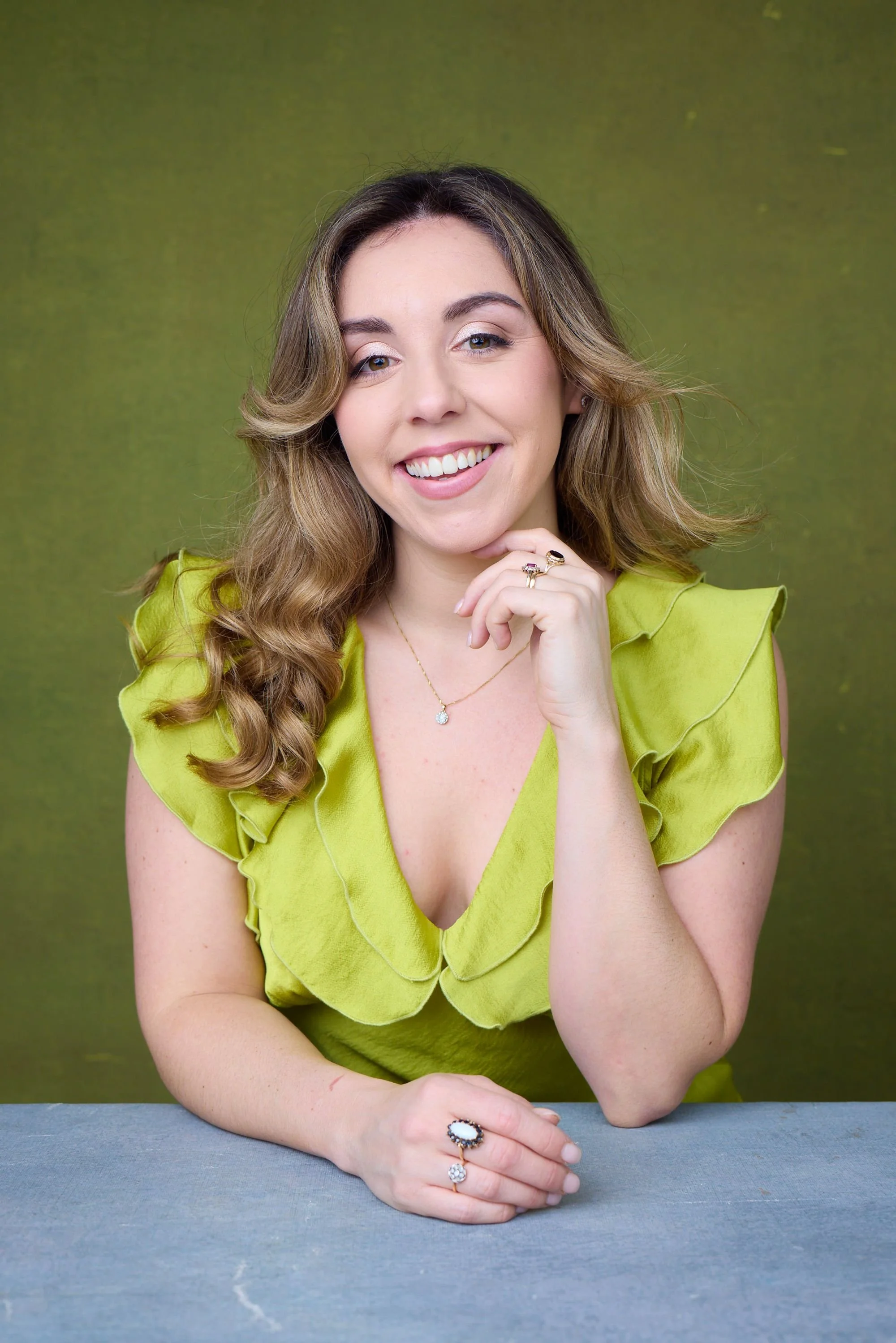 A woman with light skin and brown, wavy hair, wearing a bright green ruffled blouse, sitting at a table with a green background. She is smiling, resting her chin on her right hand, and wearing rings and a necklace.